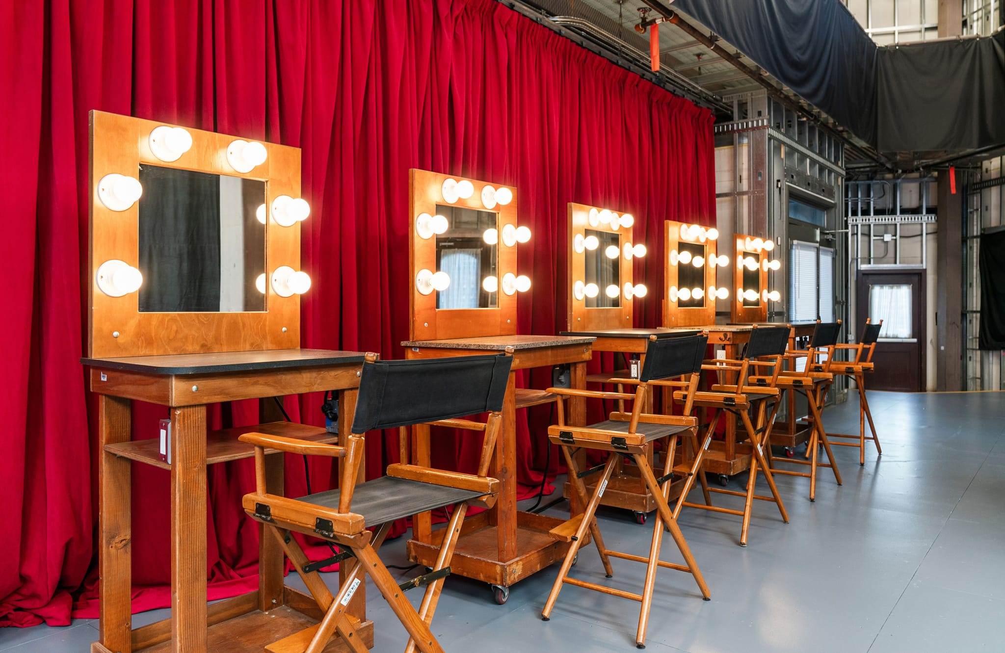 A row of makeup stations with illuminated mirrors and director's chairs against red curtains in a studio.