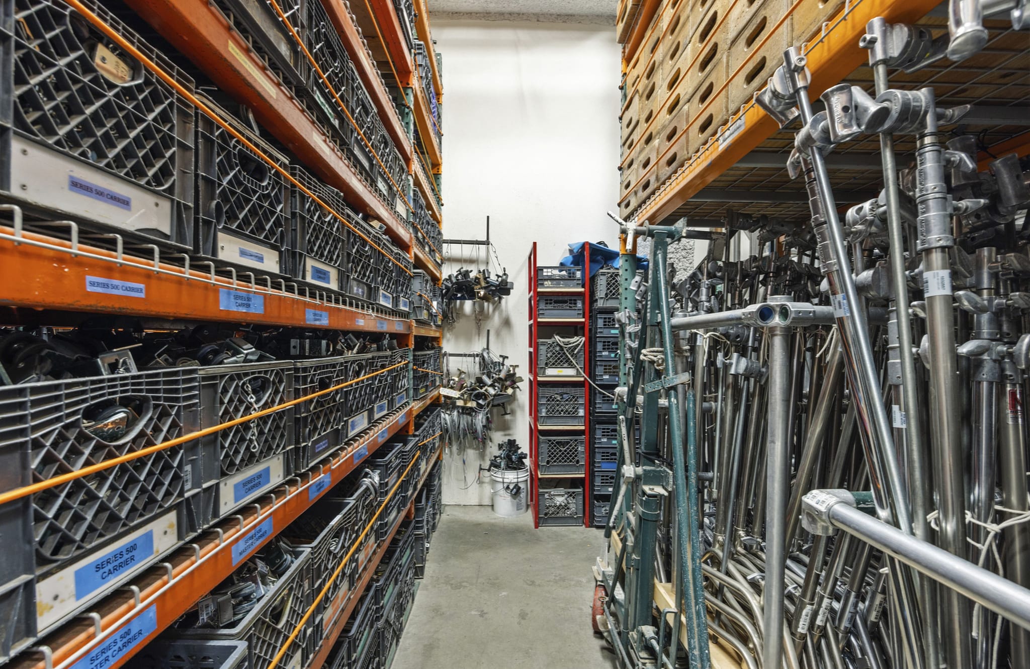 Warehouse aisle with shelves of labeled crates on the left and racks of metal stands on the right.