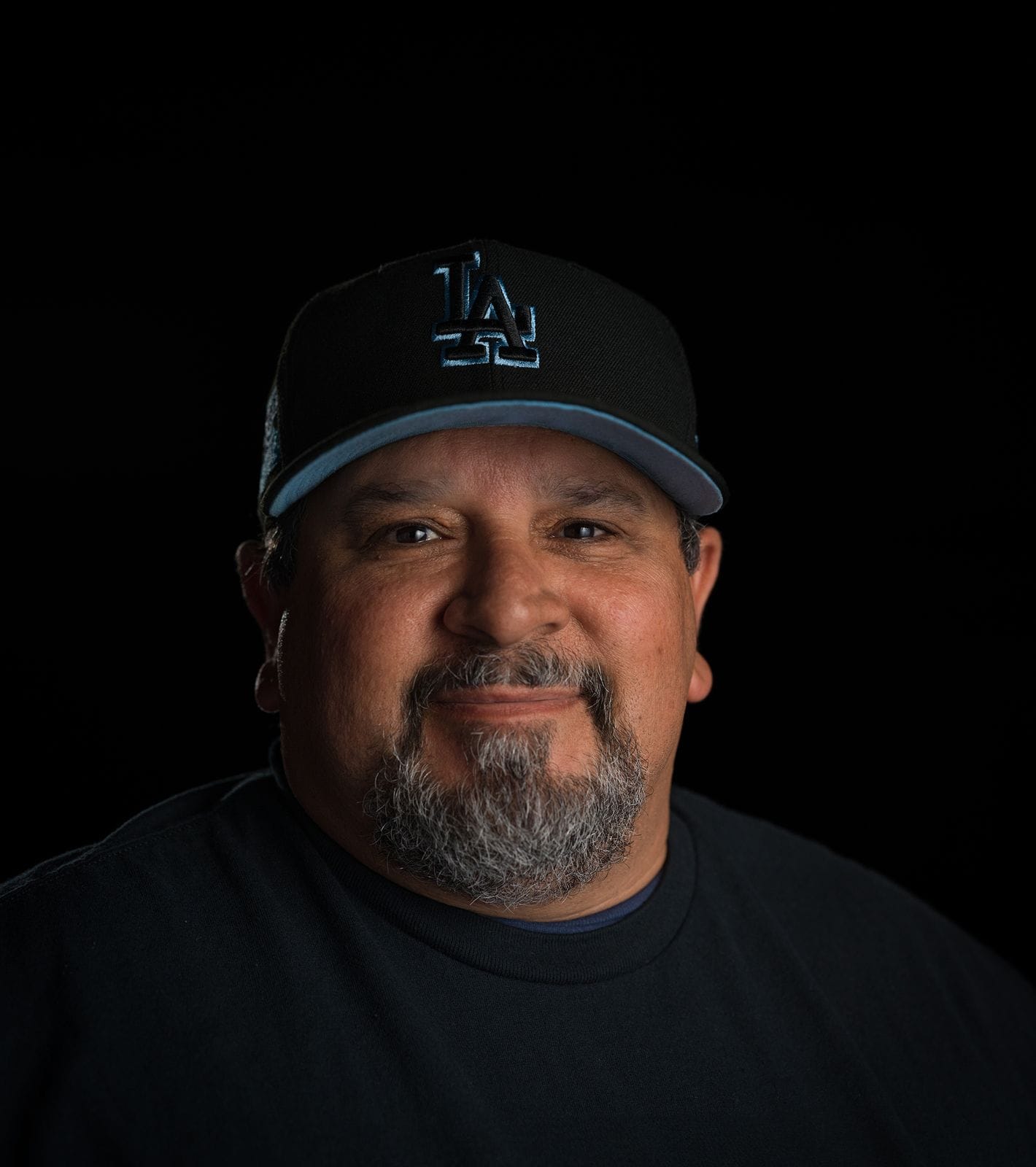 Headshot of a smiling man with a gray beard and LA Dodgers cap against a black background.