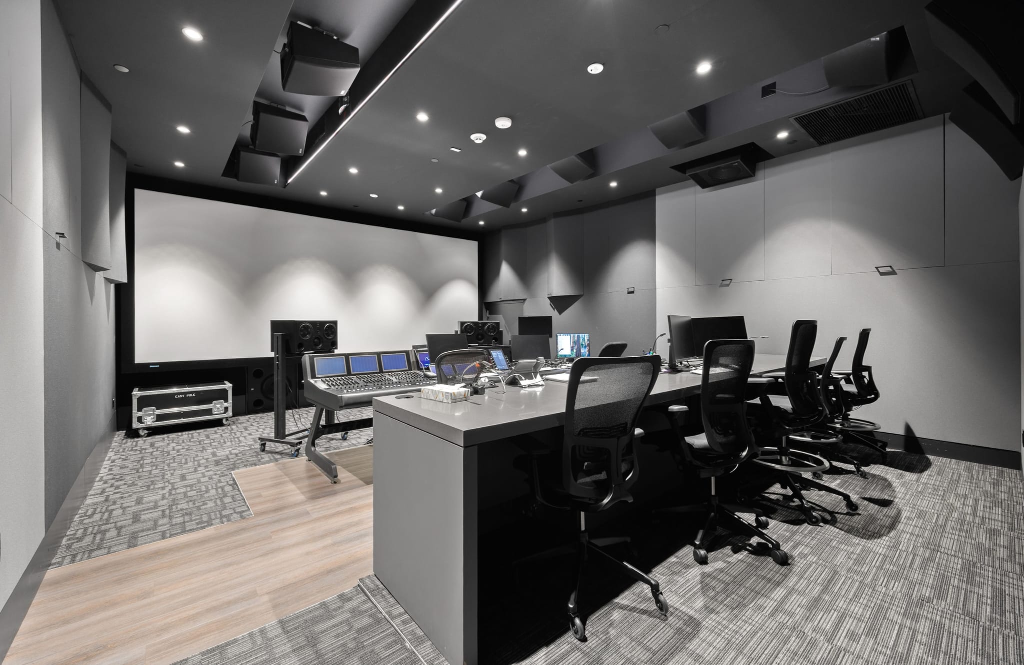 A modern, dark grey media production studio with a large projection screen, control console, and multiple workstations.