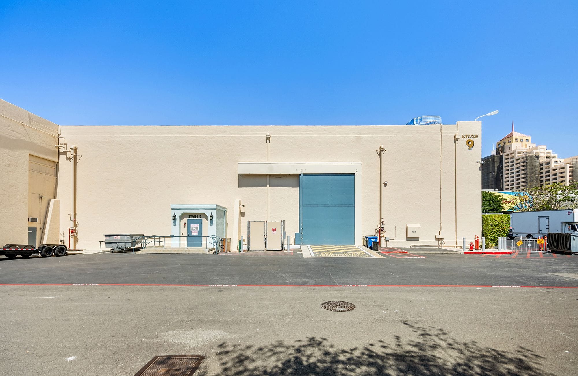 Exterior of Studio 9, a large beige building with a blue sliding door, a parking lot in the foreground, and a clear blue sky.