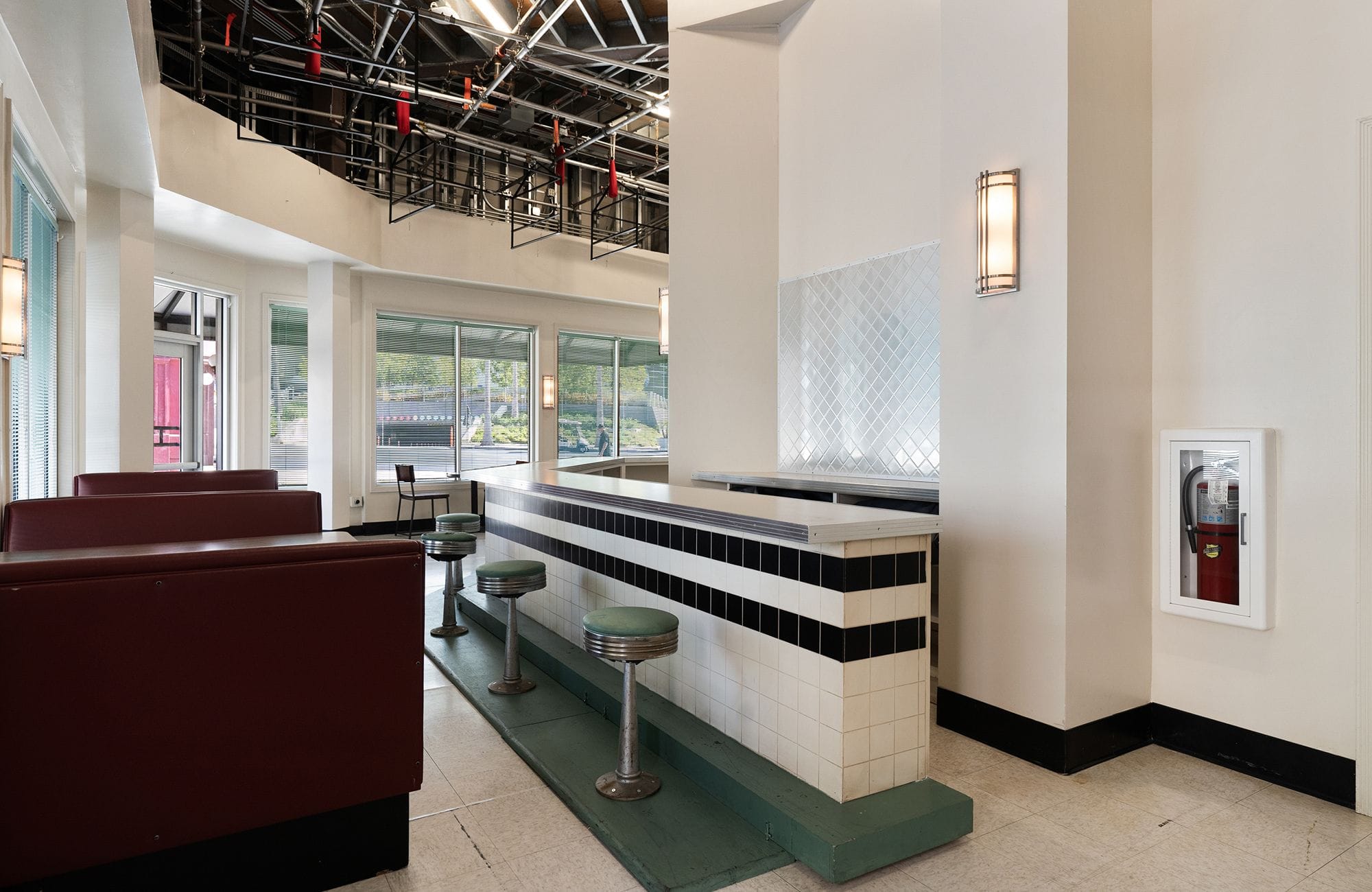 Empty retro diner with maroon booths, a tiled counter with green stools, and an exposed ceiling.