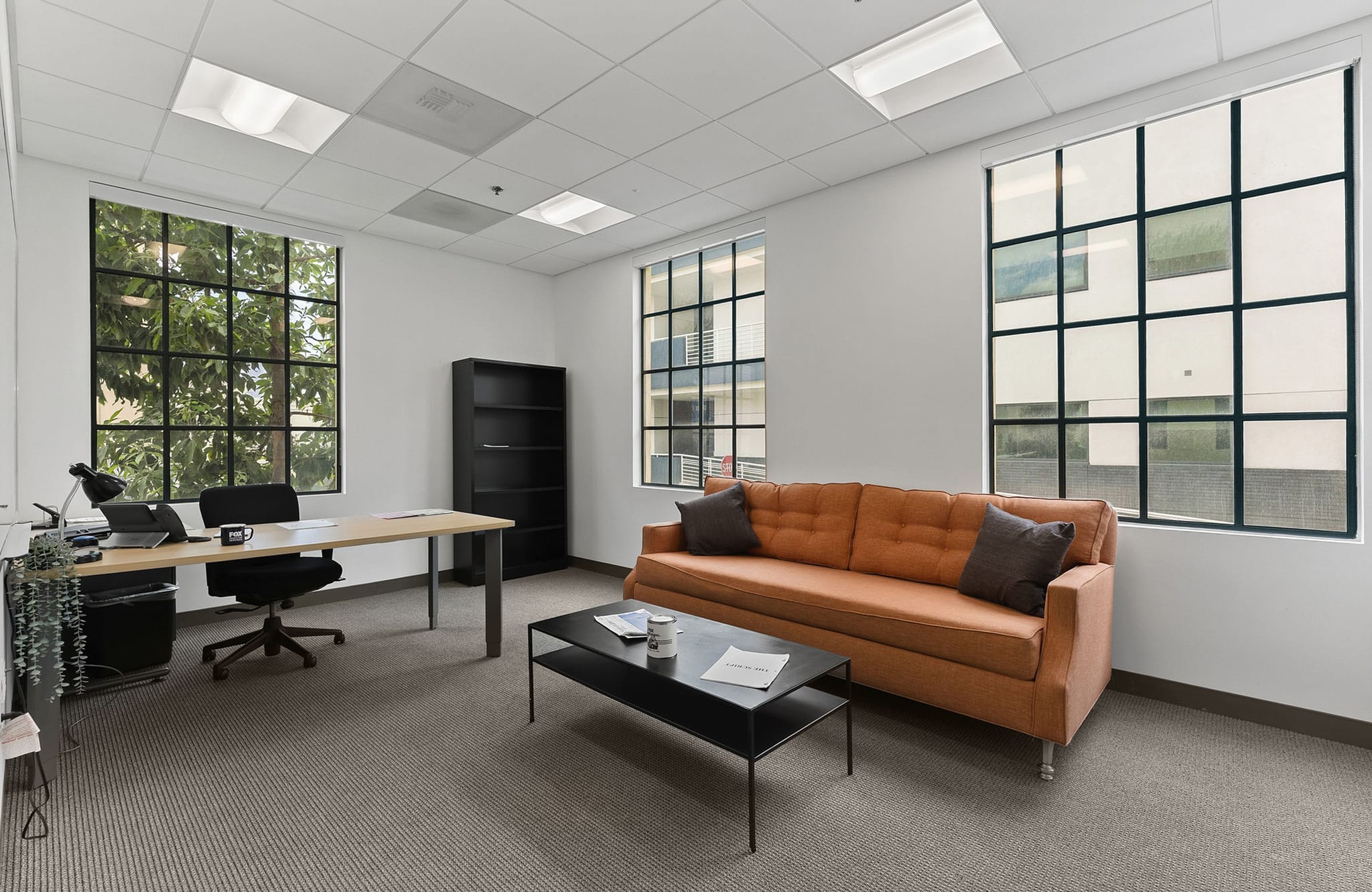 A bright office room with a desk and chair, an orange sofa and coffee table, and large grid-patterned windows.