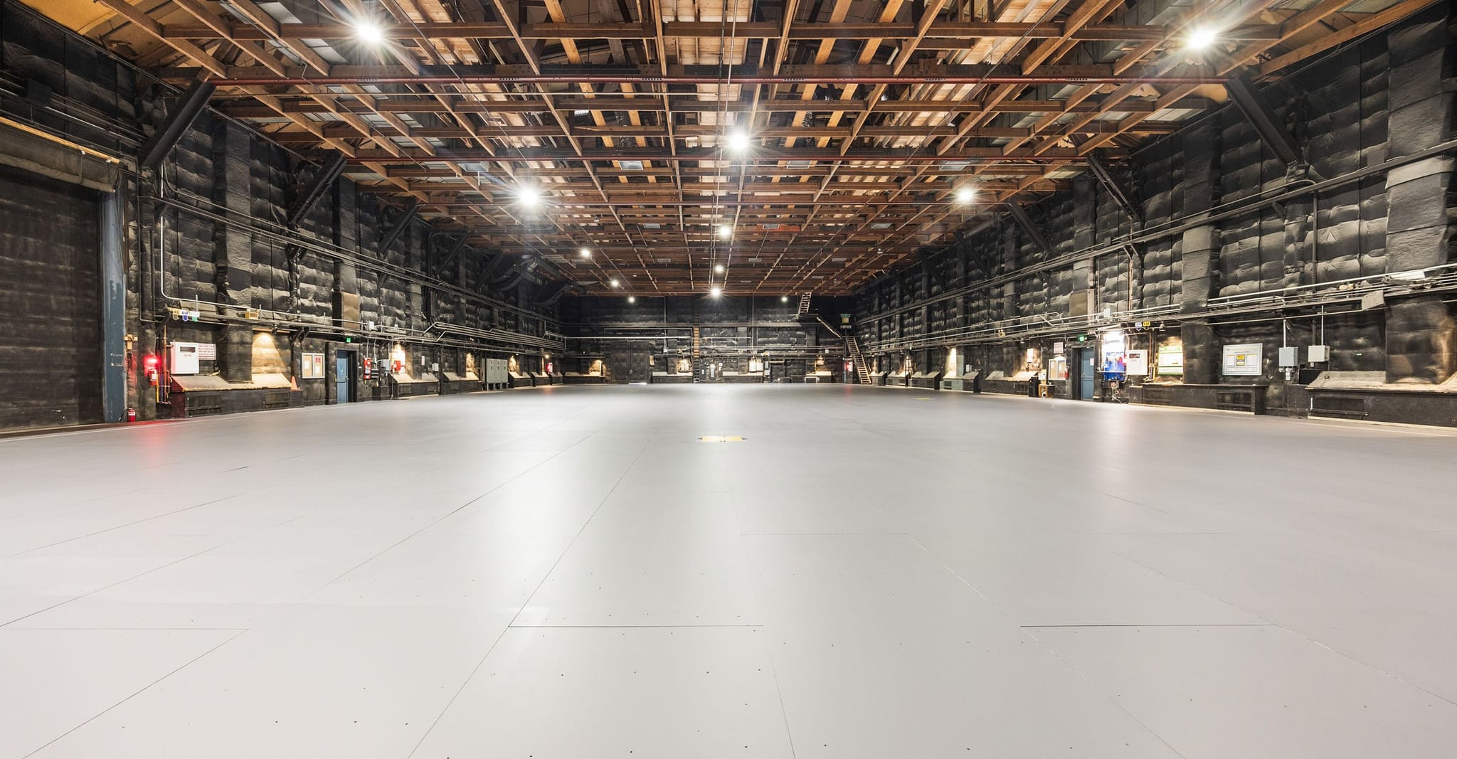 A large, empty industrial-style room with a light grey floor, dark walls, and an exposed wooden beam ceiling.