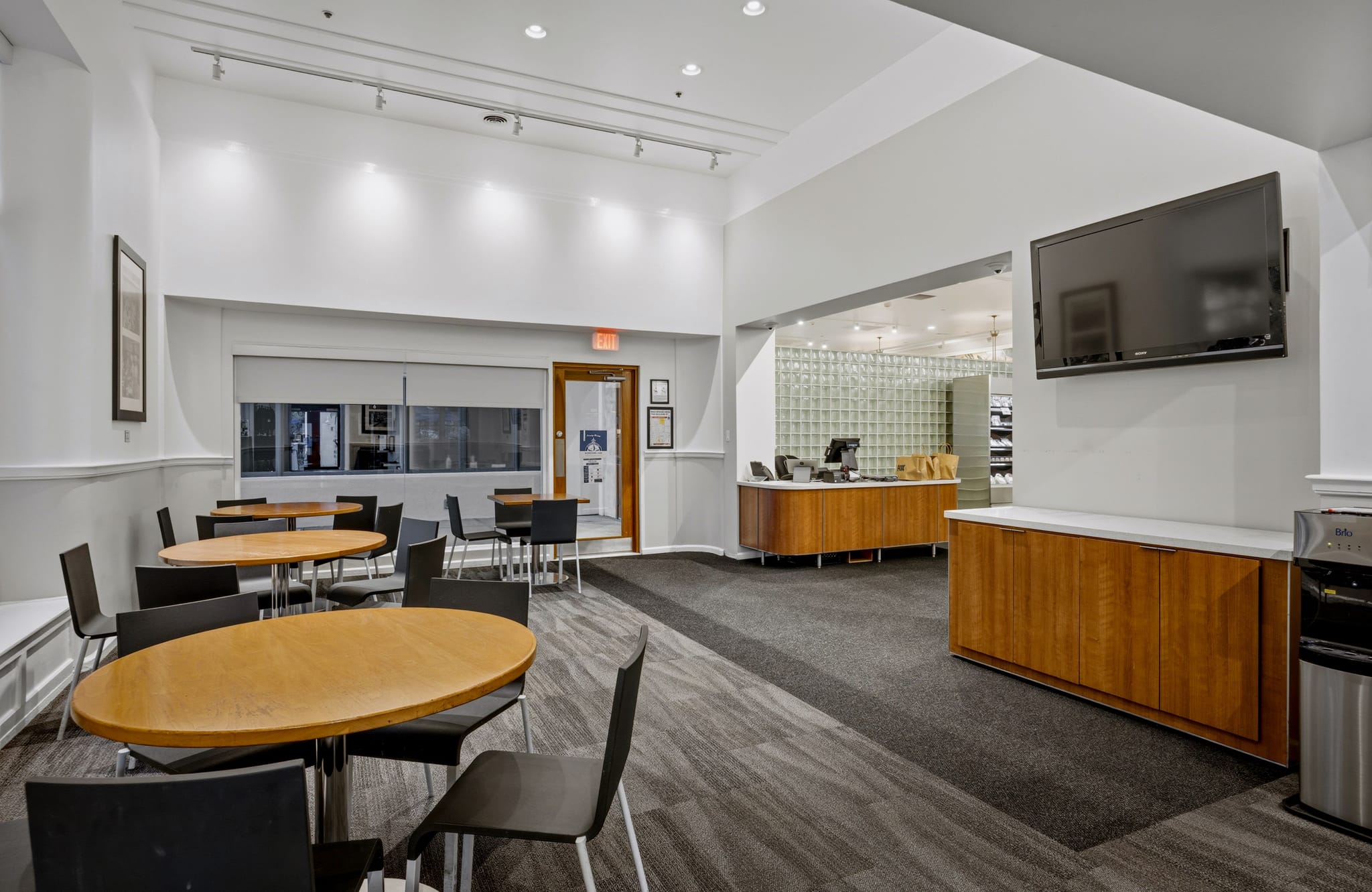 A bright, modern lounge with several wooden tables and black chairs, a long wooden reception desk, and a wall-mounted TV.
