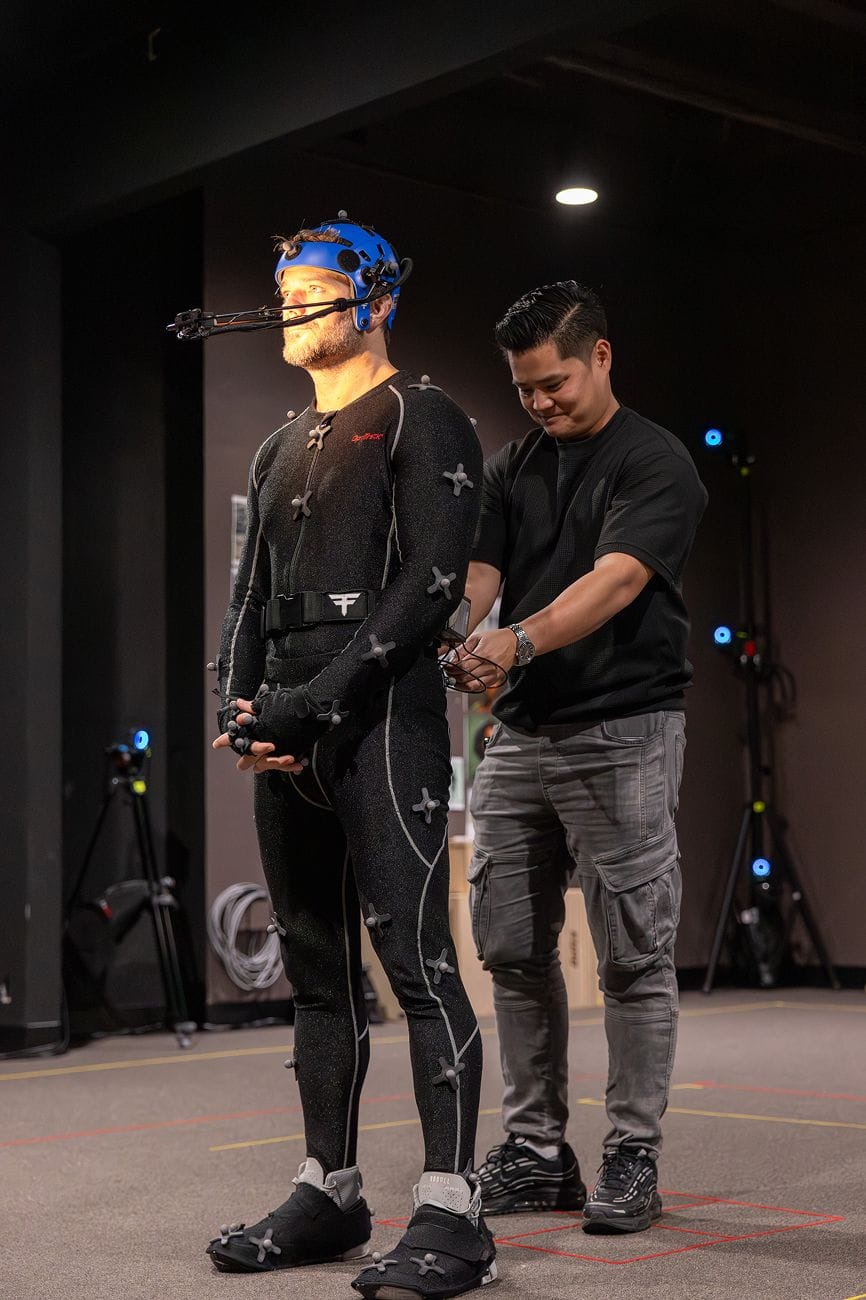 A man in a black motion capture suit and facial capture helmet is adjusted by another man in a dark studio.