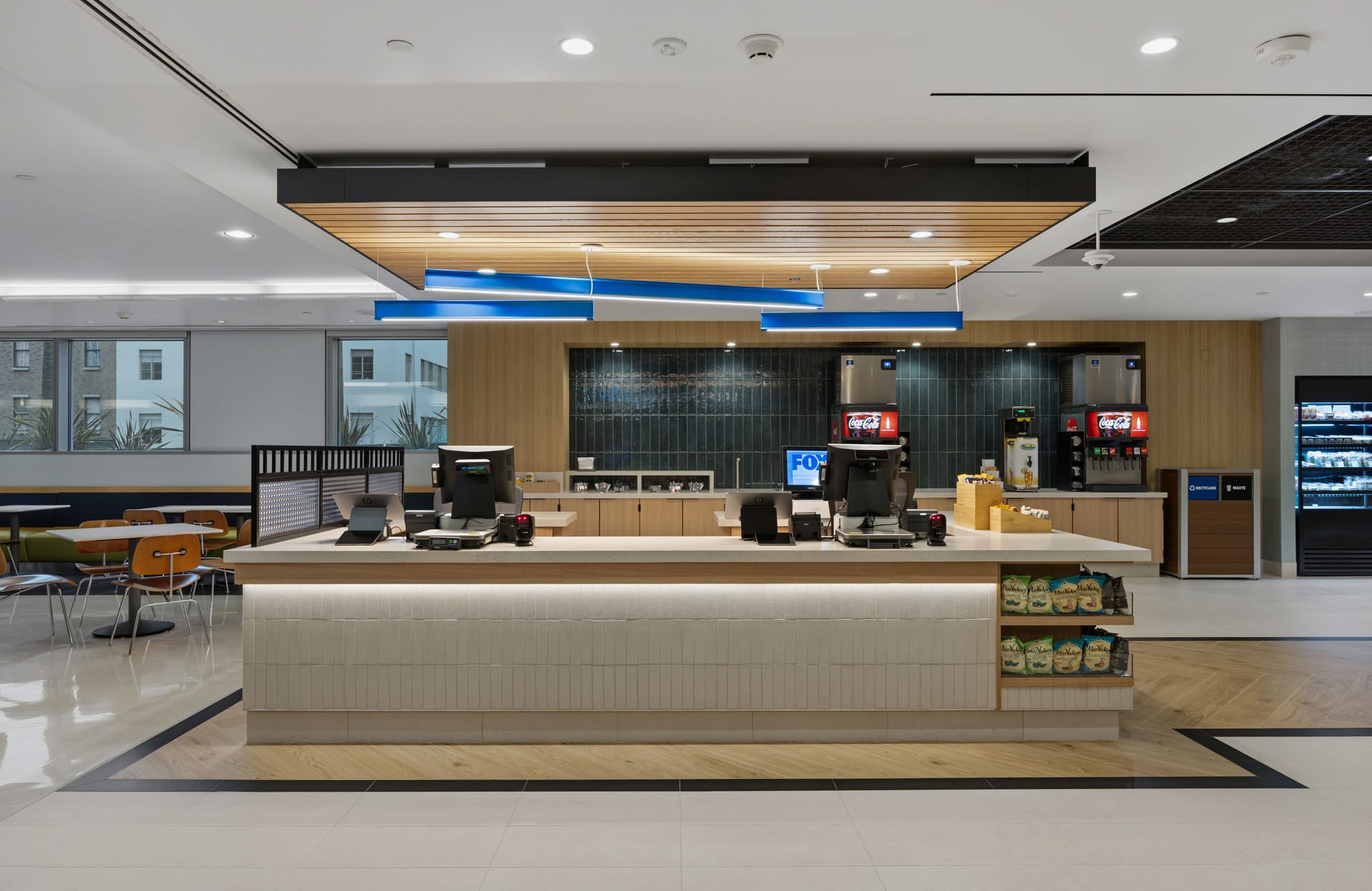 A bright, modern self-service cafeteria with a long counter, drink machines, a grab-and-go cooler, and a seating area.