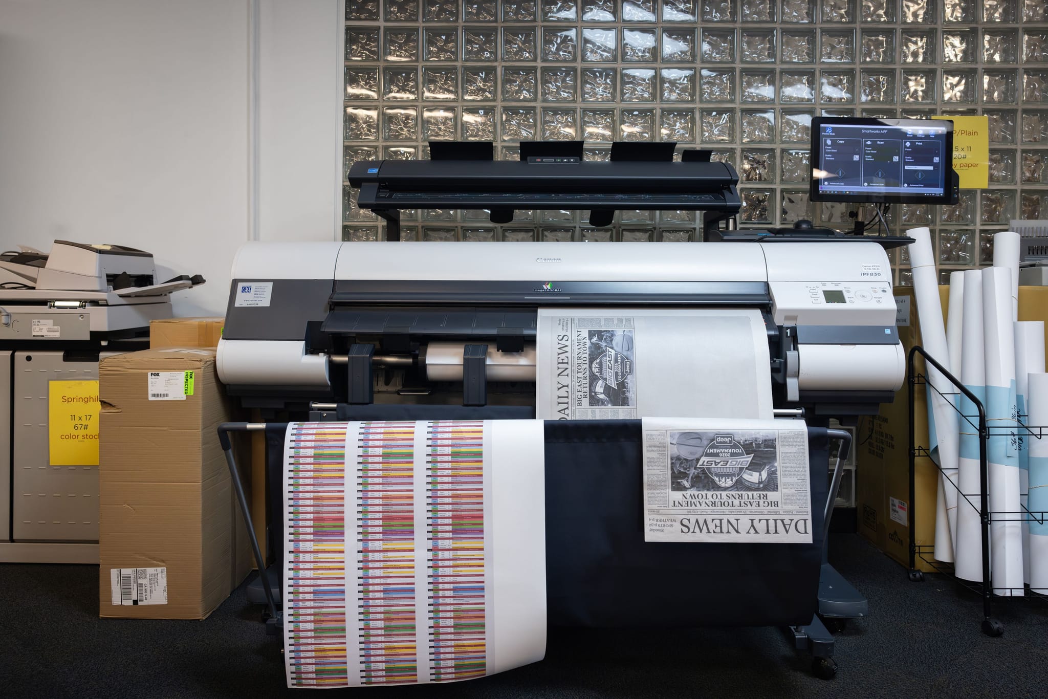A large-format printer in a print shop, displaying a "Daily News" newspaper print and a colorful striped test pattern on its output rolls.