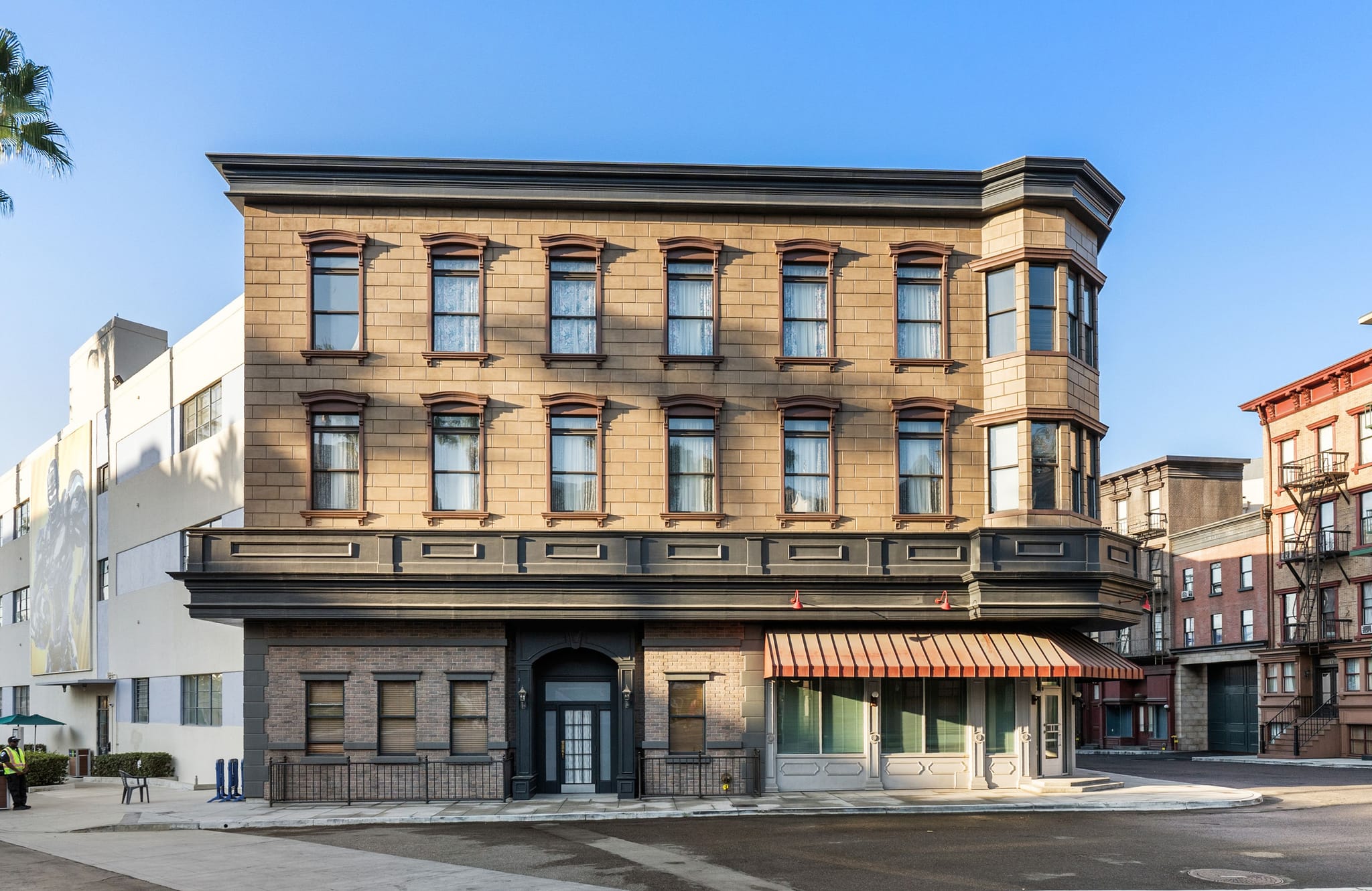 An old-fashioned, tan multi-story building with a bay window and storefront awning, on a street scene likely a film set.