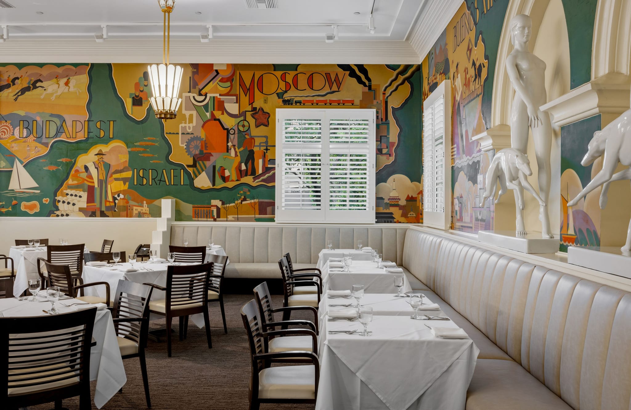 A restaurant dining room with a large, colorful Art Deco travel mural, white booth seating, tables with white tablecloths, and white classical statues on a ledge.