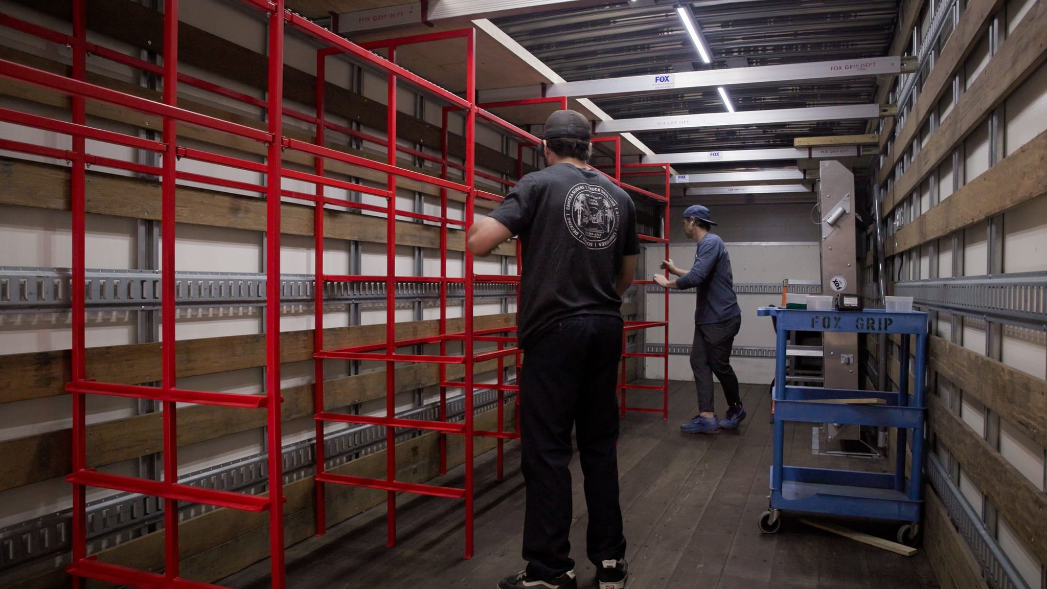 Two men work on installing bright red shelving inside a wood-lined truck trailer.