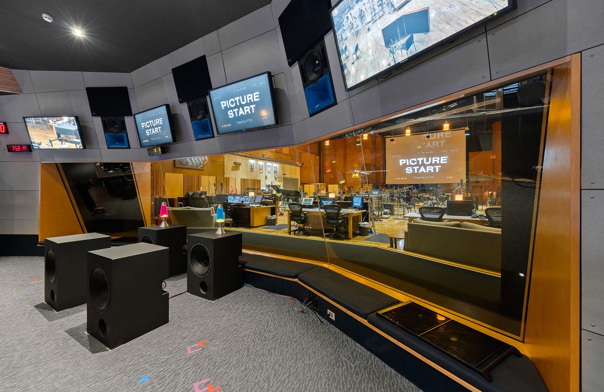 An empty post-production studio with subwoofers in the viewing area and a control room visible through glass, with multiple screens displaying "PICTURE START".