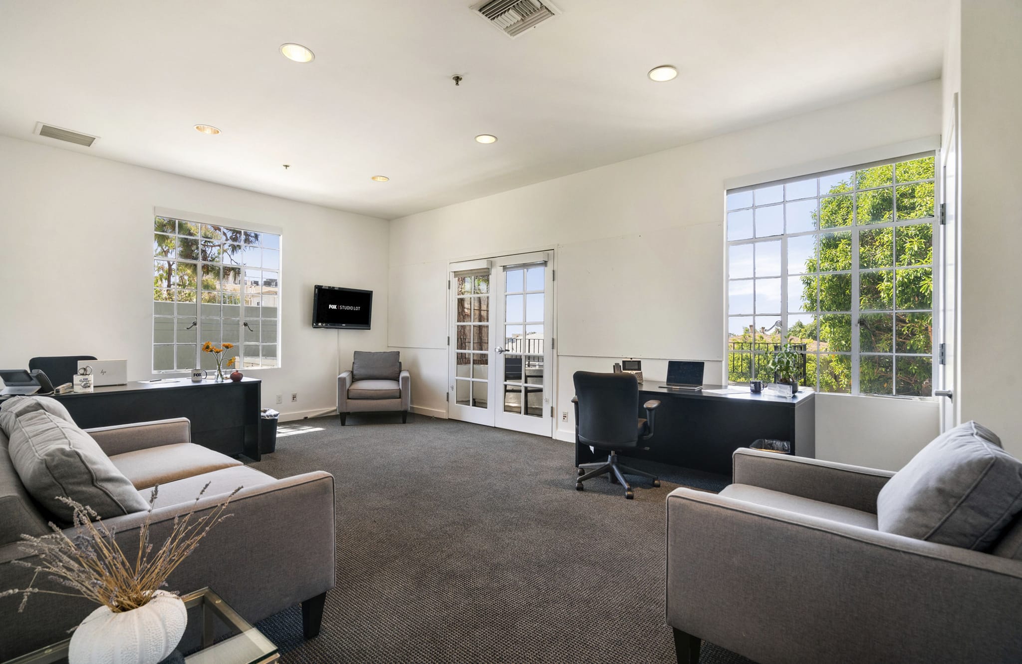 A modern office or common room with two desks, a grey sofa, armchairs, and large windows.