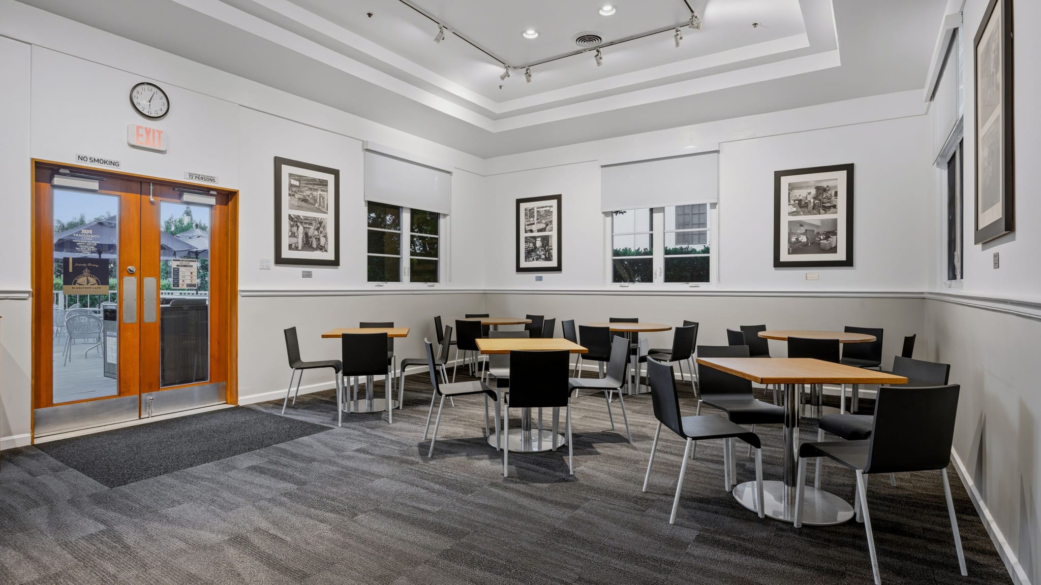 A bright, empty room with small cafe tables and chairs, framed art on white walls, and a glass double door.