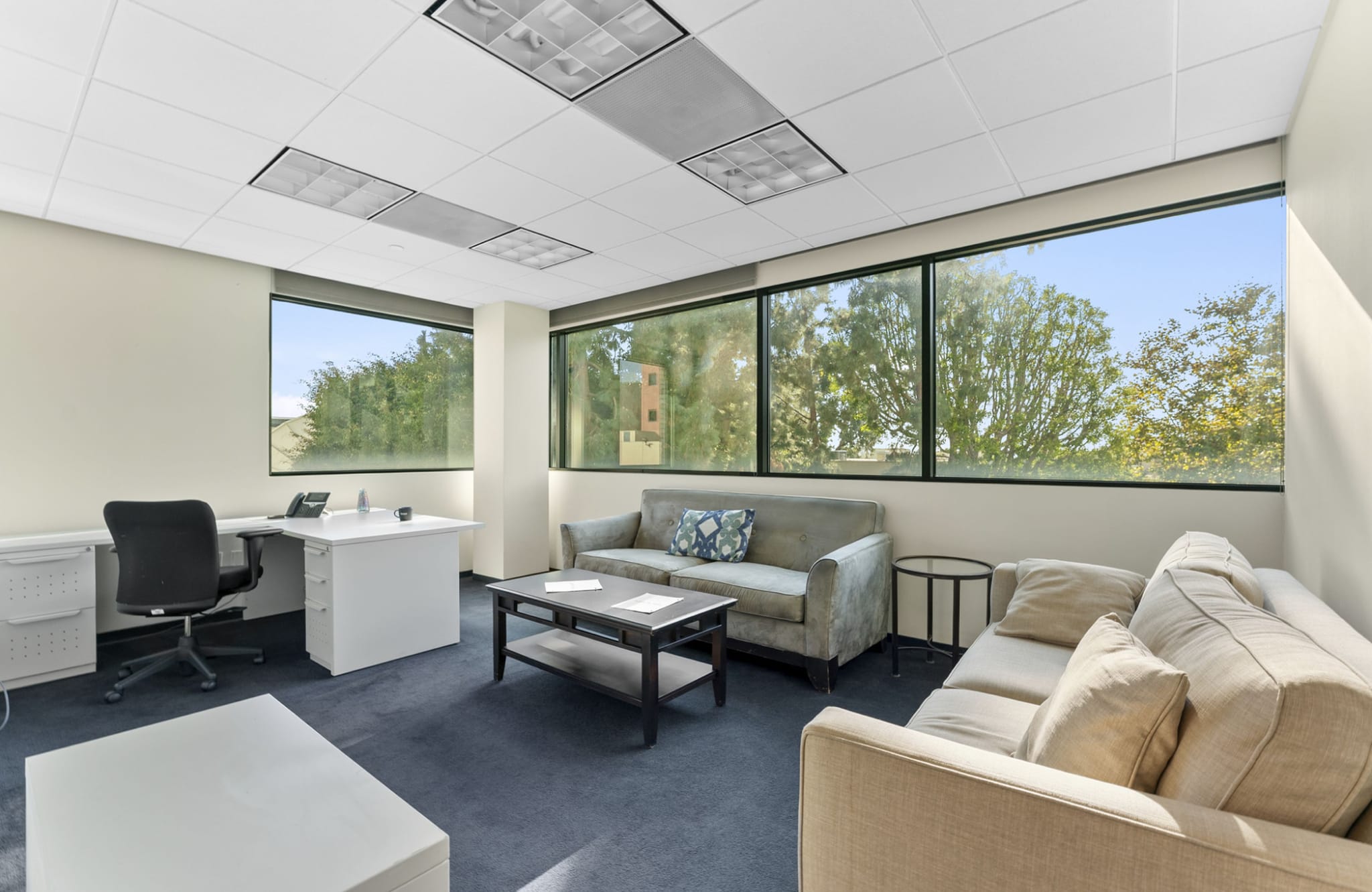 A modern office space with a desk and chair, two sofas, and large windows overlooking trees.