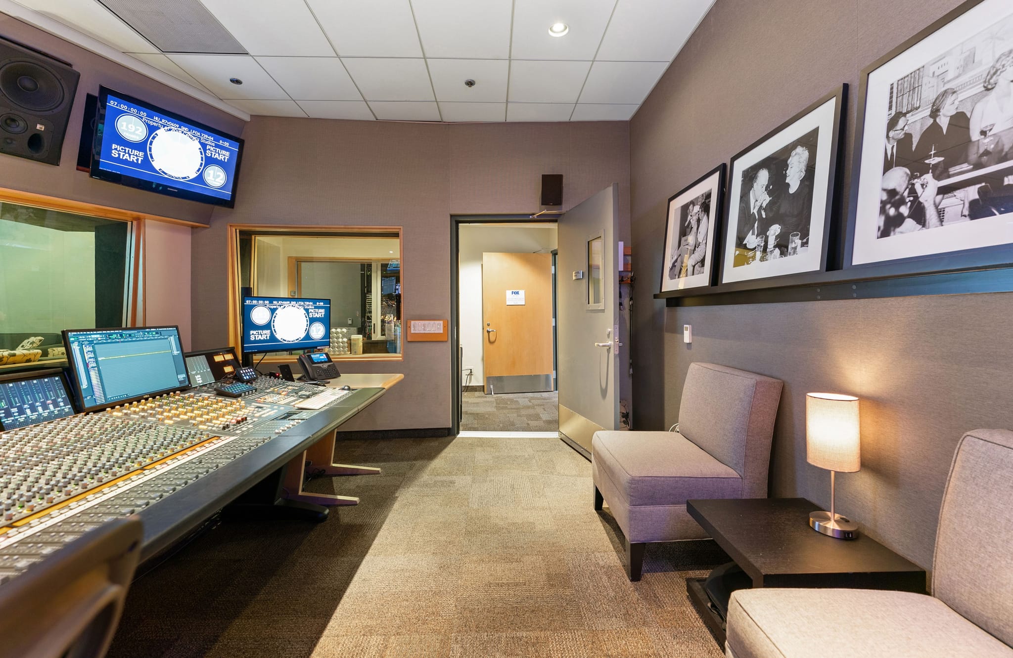 A recording studio control room with a large mixing console, computer monitors, and a lounge area with framed photos.