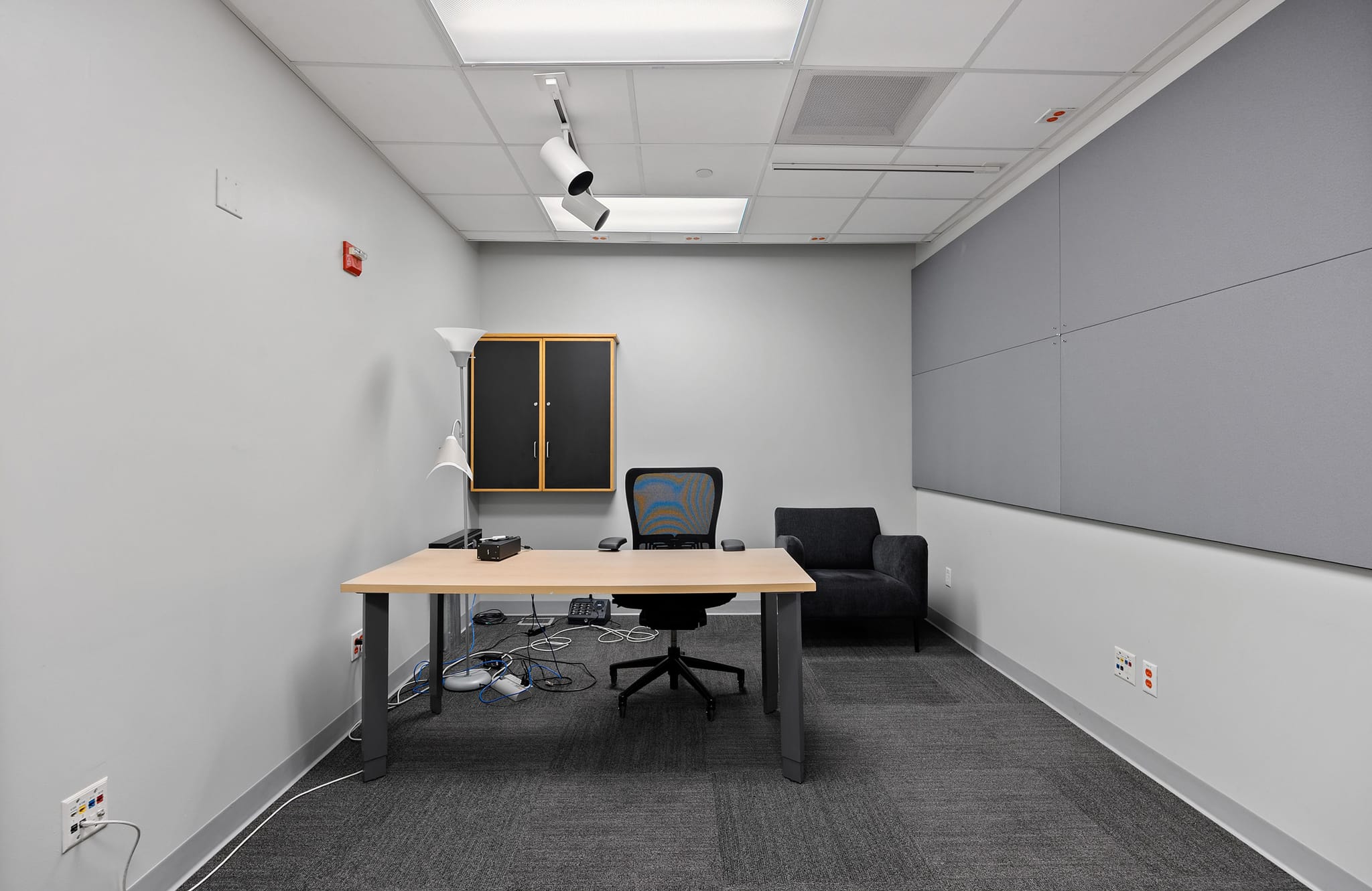 A modern office room with a light wooden desk, a black office chair, a black armchair, and grey acoustic panels on one wall.