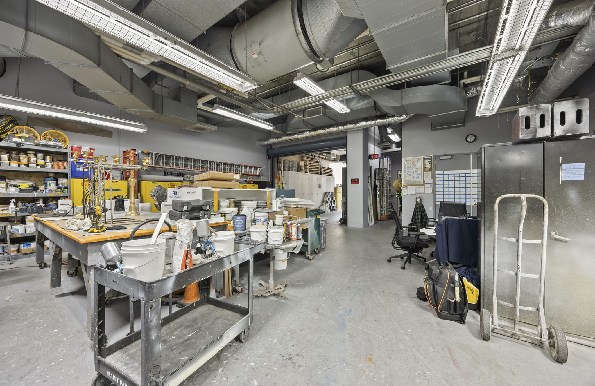 A spacious industrial workshop with exposed ductwork, workbenches full of paint and tools, and a desk area next to metal storage cabinets and a hand truck.