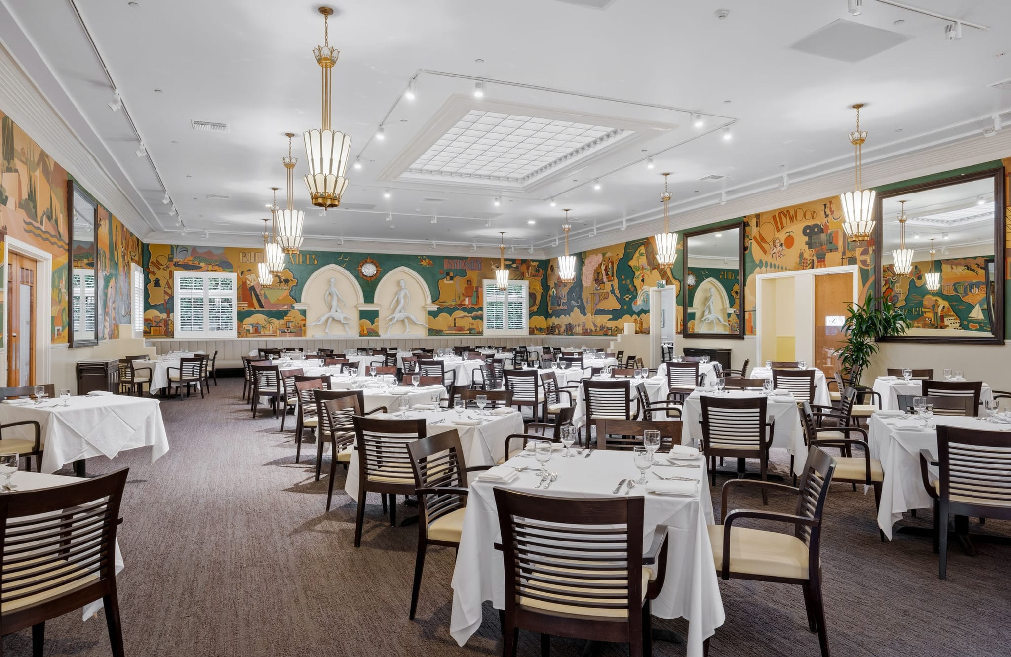 An elegant dining room with white-clothed tables, colorful murals, ornate ceiling lights, and large mirrors.