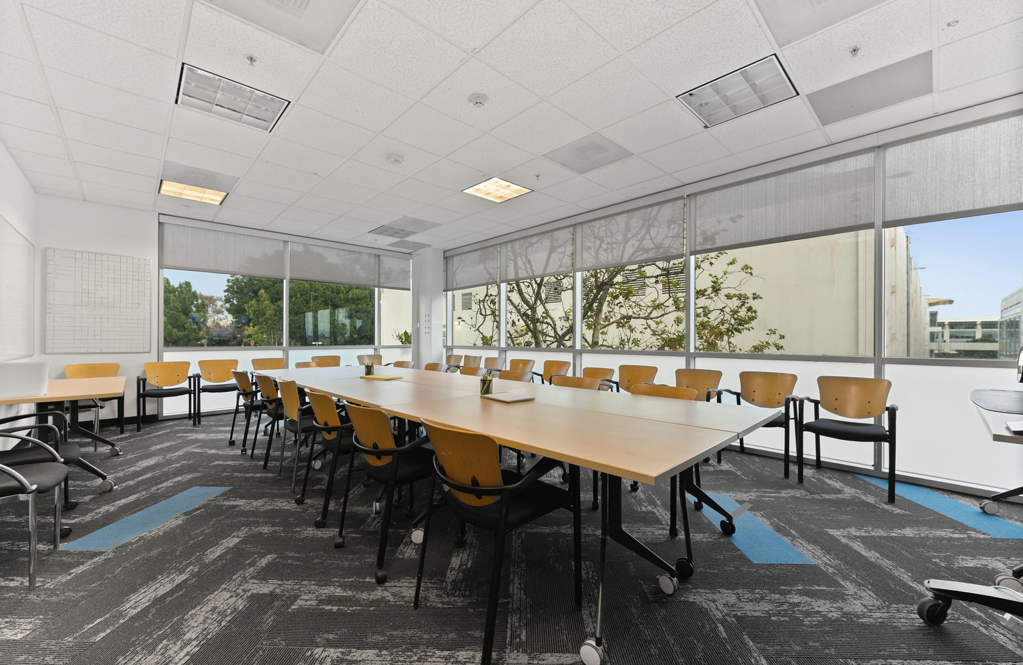 A modern conference room with a long wooden table, multiple chairs, large windows with blinds, and patterned carpet.