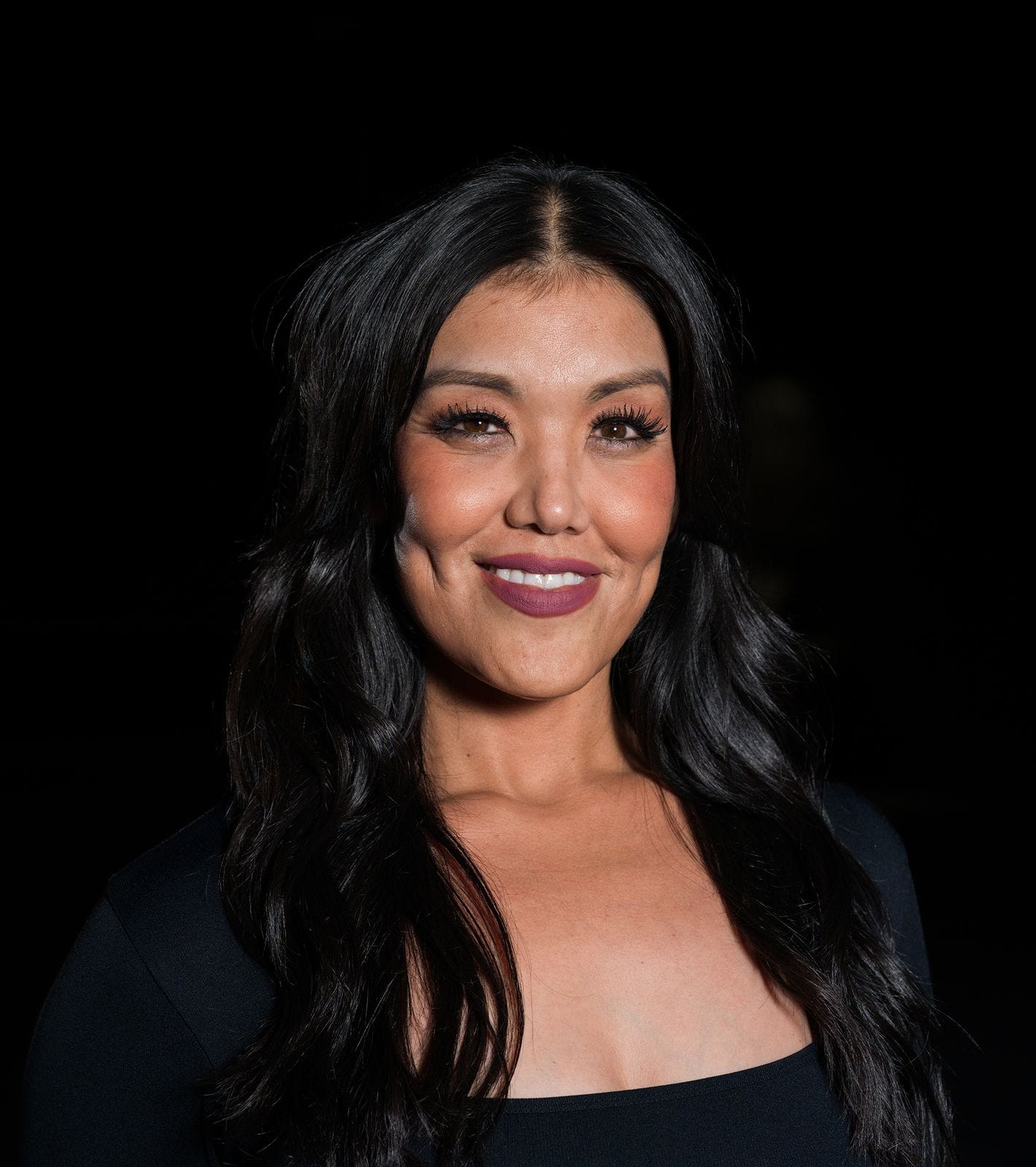A smiling woman with long dark hair, dimples, and dark lipstick, wearing a black top against a dark background.