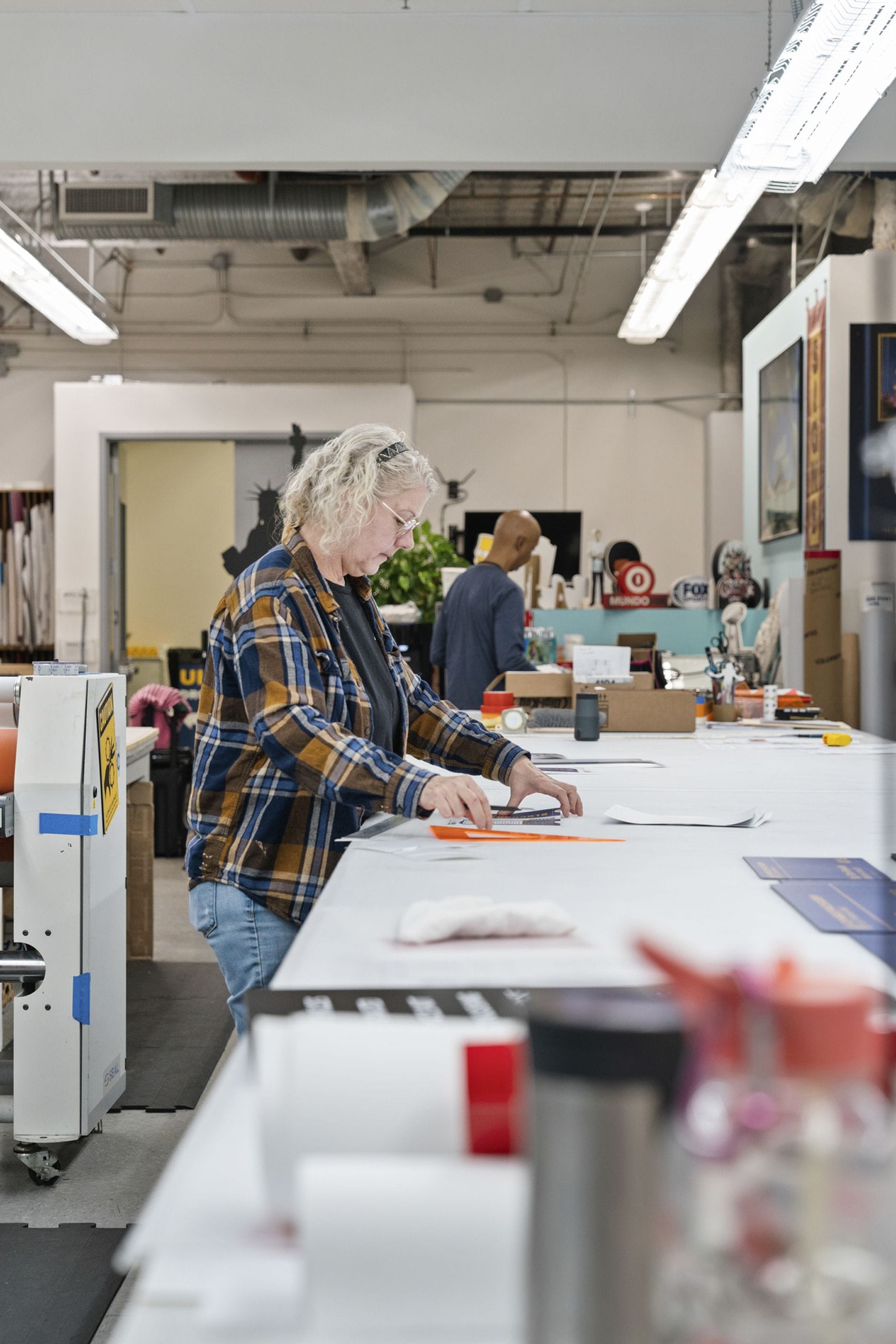 A woman in a plaid shirt and glasses works with paper at a large table in a brightly lit workshop.