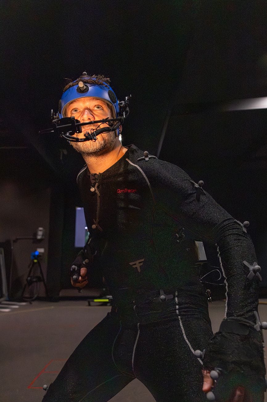A man in a motion capture suit and helmet with a facial capture rig looks up intently in a dark studio.