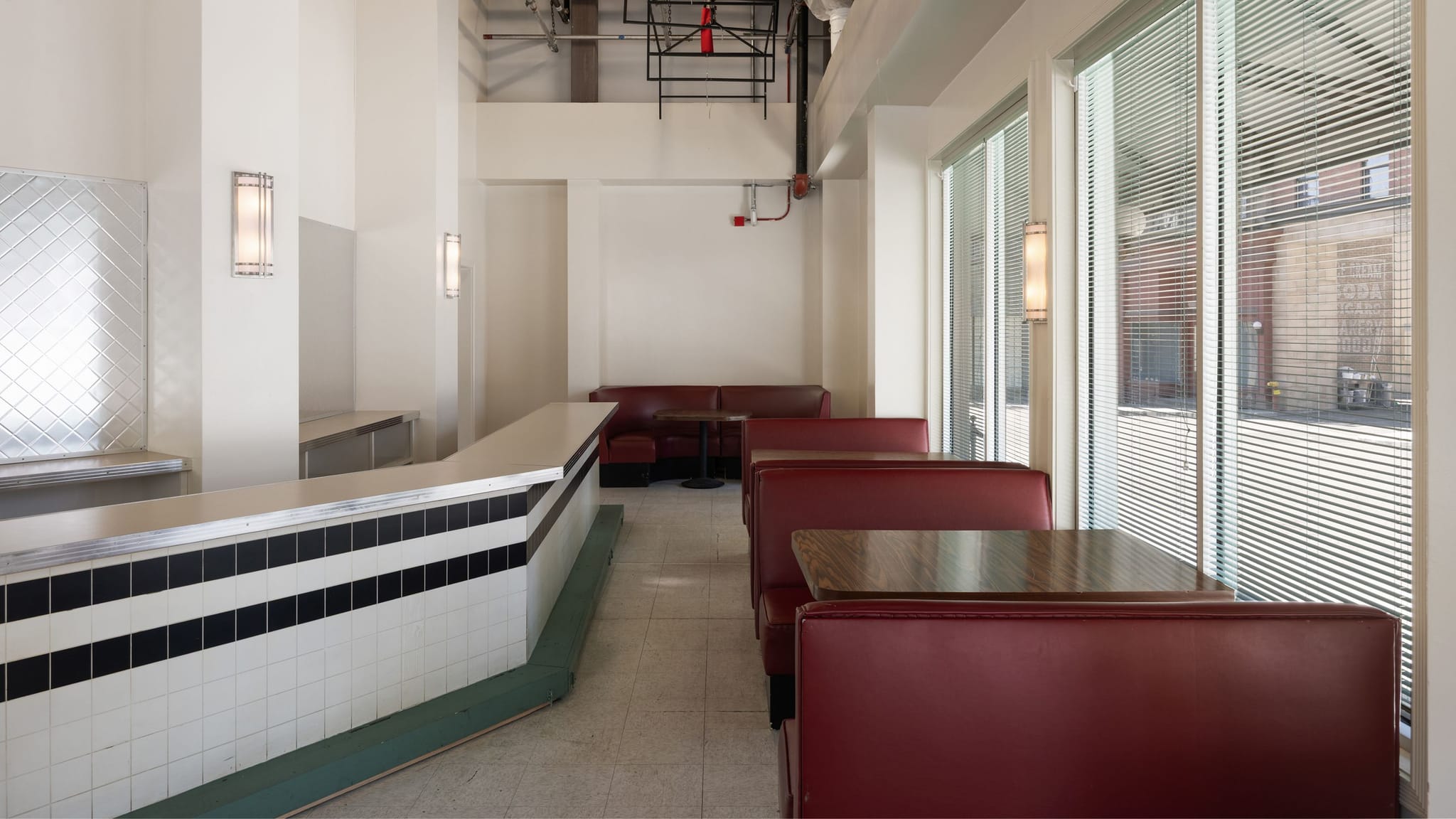 Empty retro diner interior with a white and black tiled counter, red booths, and large windows with blinds.