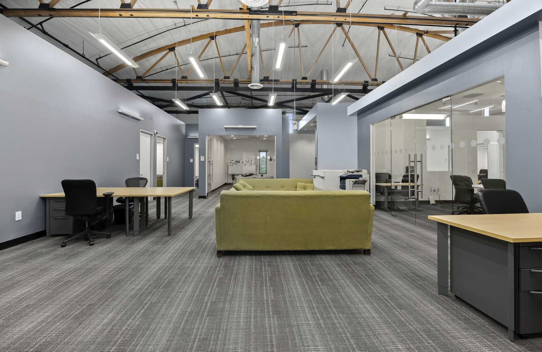 Modern open-plan office space with exposed wooden beams, a green sofa, and glass-walled private offices.