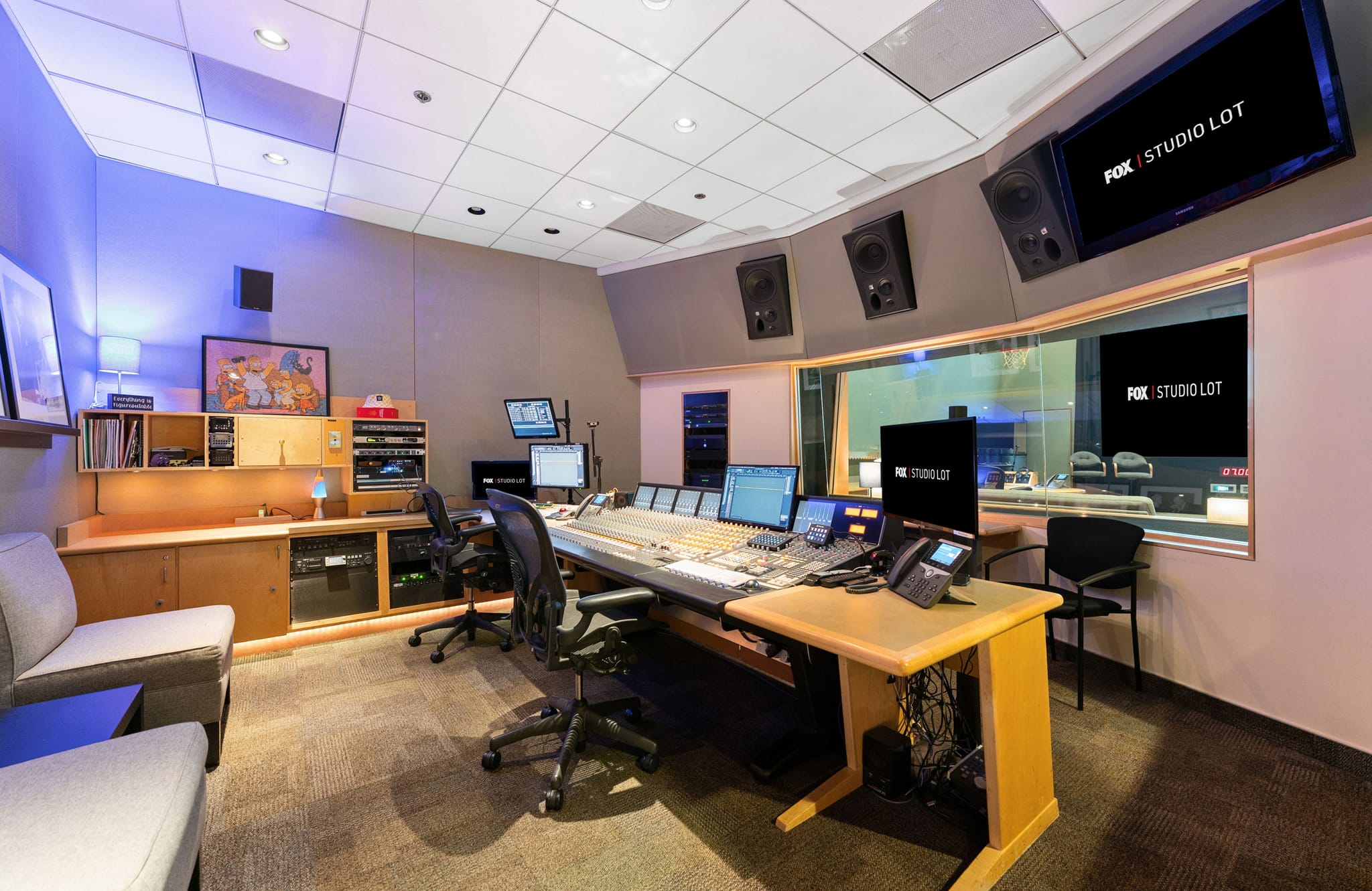 A professional audio and video control room with a large mixing console, multiple monitors displaying 'FOX | STUDIO LOT', a soundproof window into a studio, and a small lounge area.