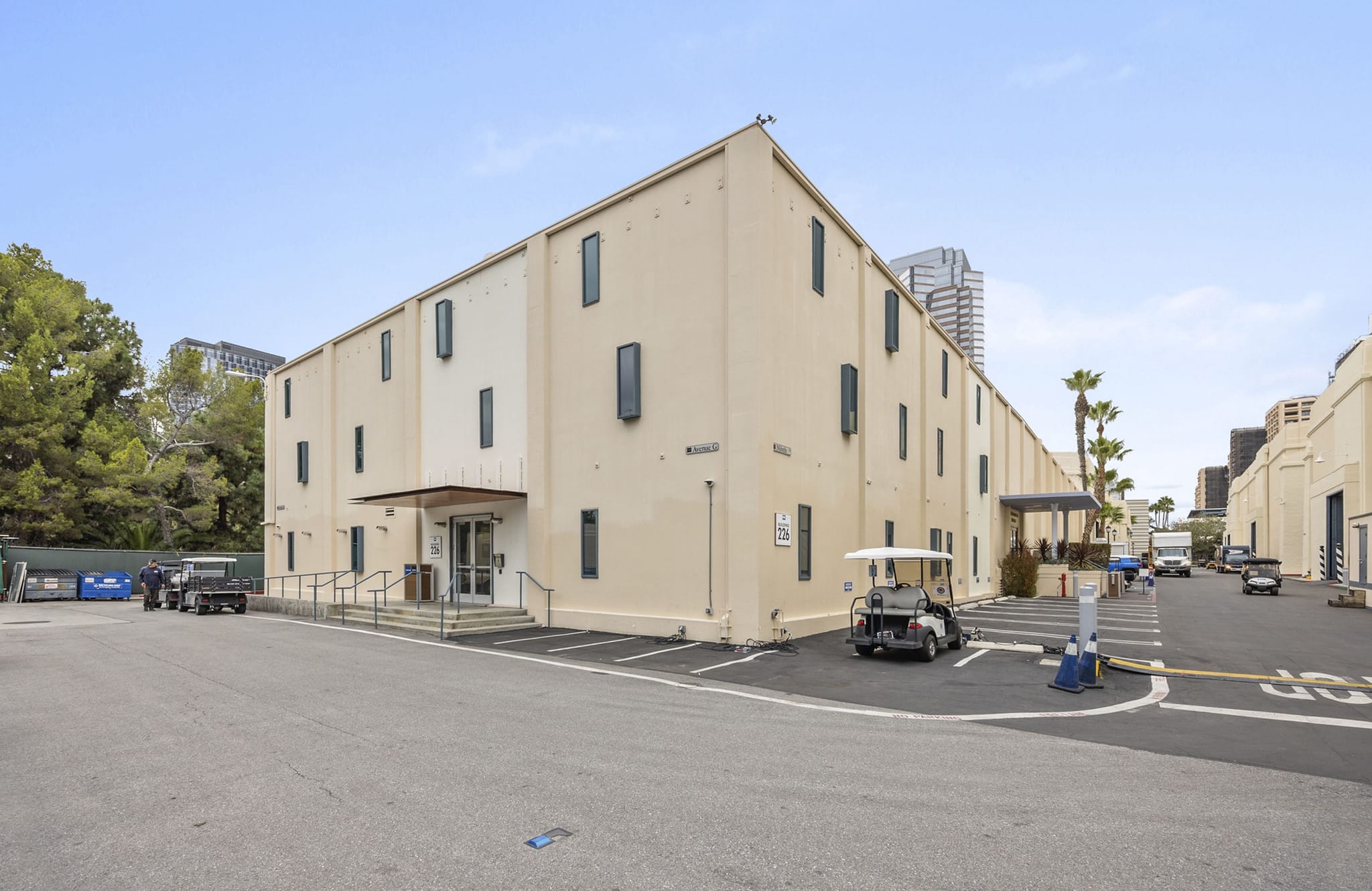 A long, cream-colored building with small windows, an asphalt lot, a golf cart, trees, and distant city buildings under a clear sky.