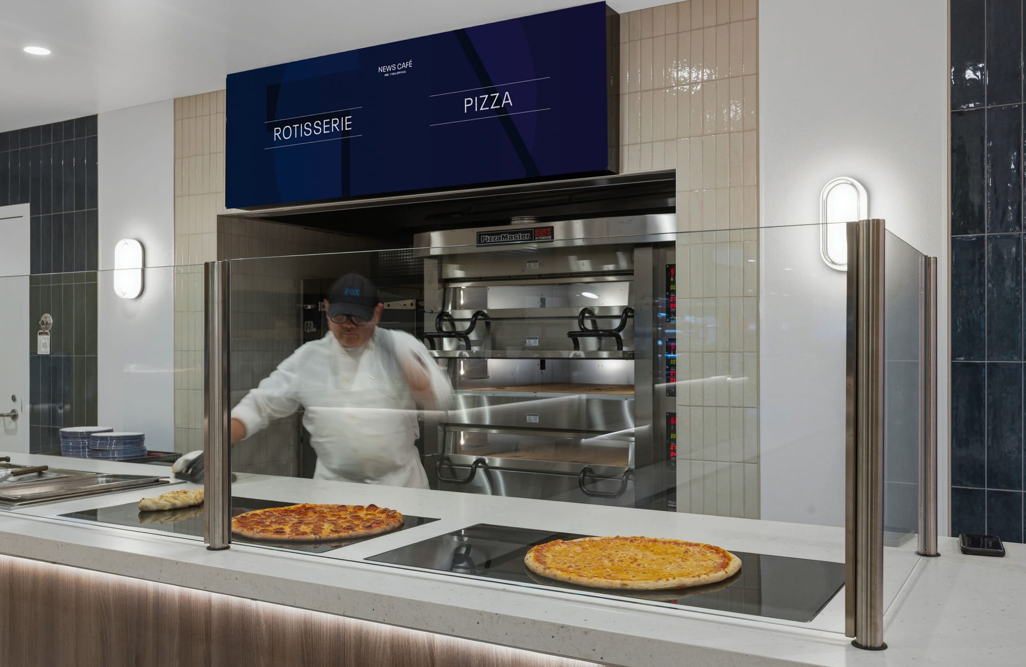 Chef behind a glass counter with two pizzas on warmers in a modern kitchen, beneath a digital menu board displaying "Rotisserie" and "Pizza."