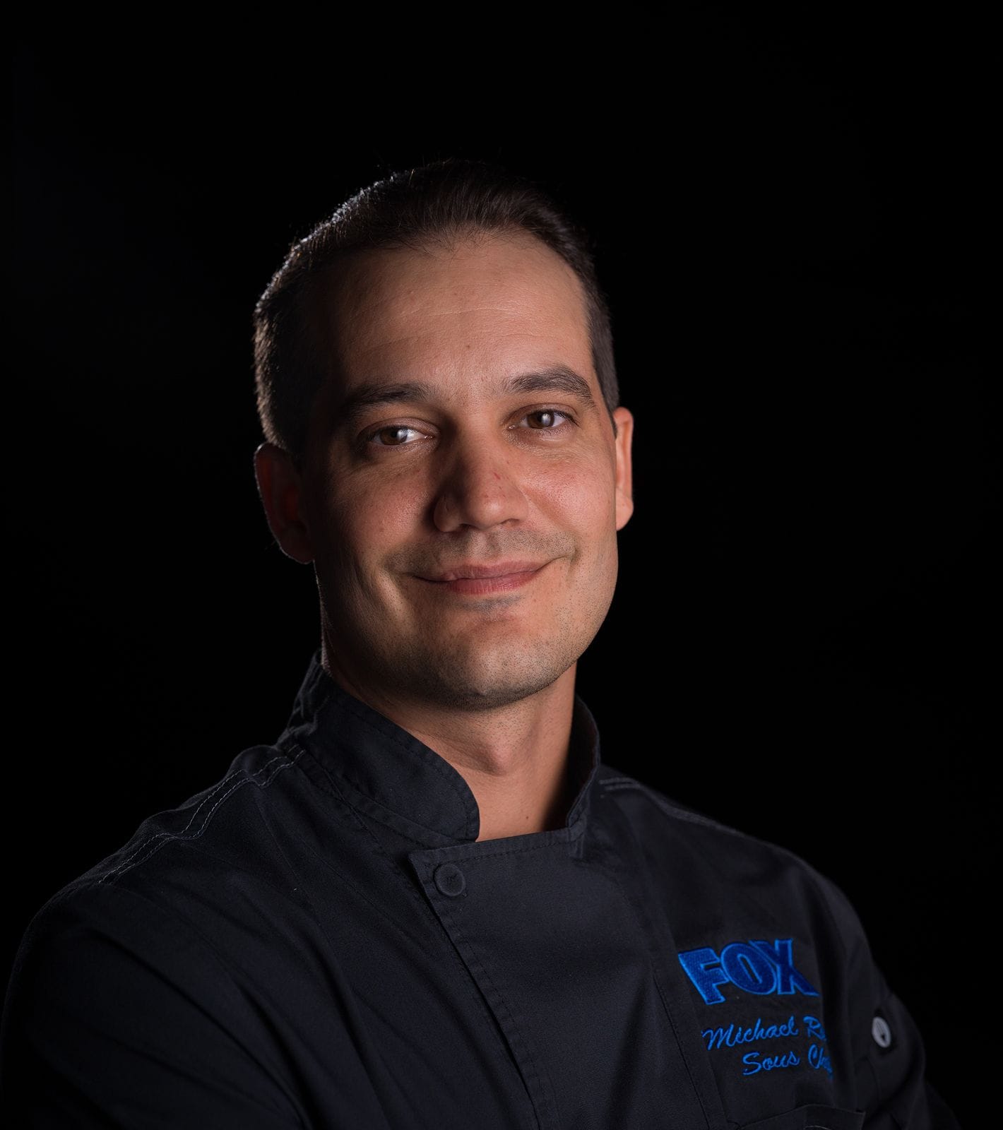 Smiling man in a black chef's jacket with a FOX logo and "Sous Chef" embroidered on the chest.