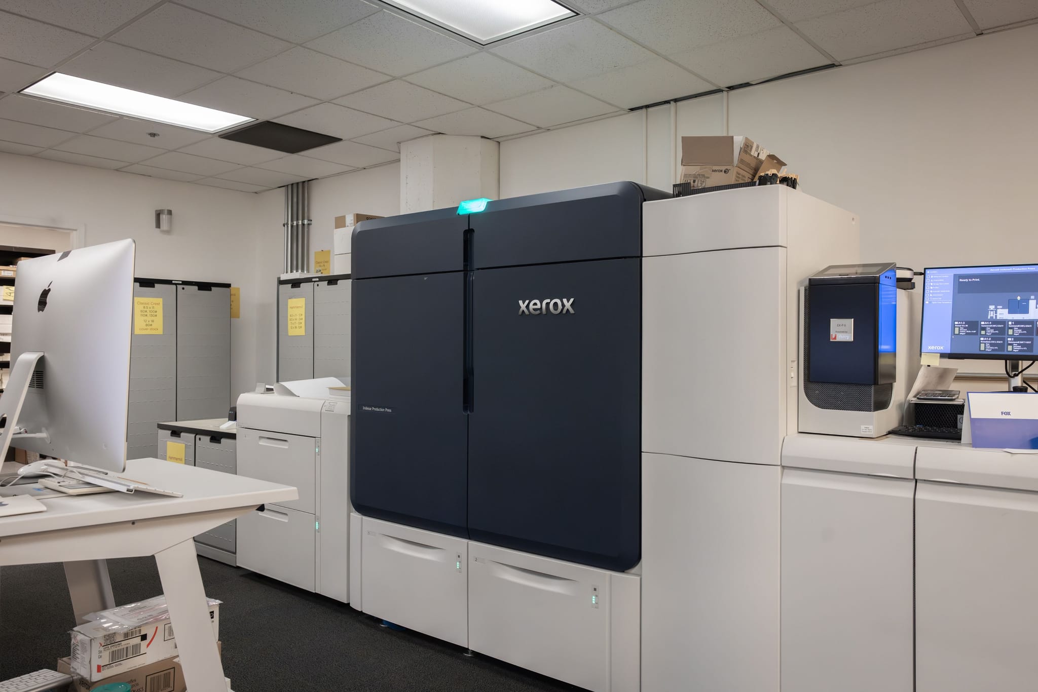 A large dark blue and white Xerox digital press in a professional printing room with a computer workstation.