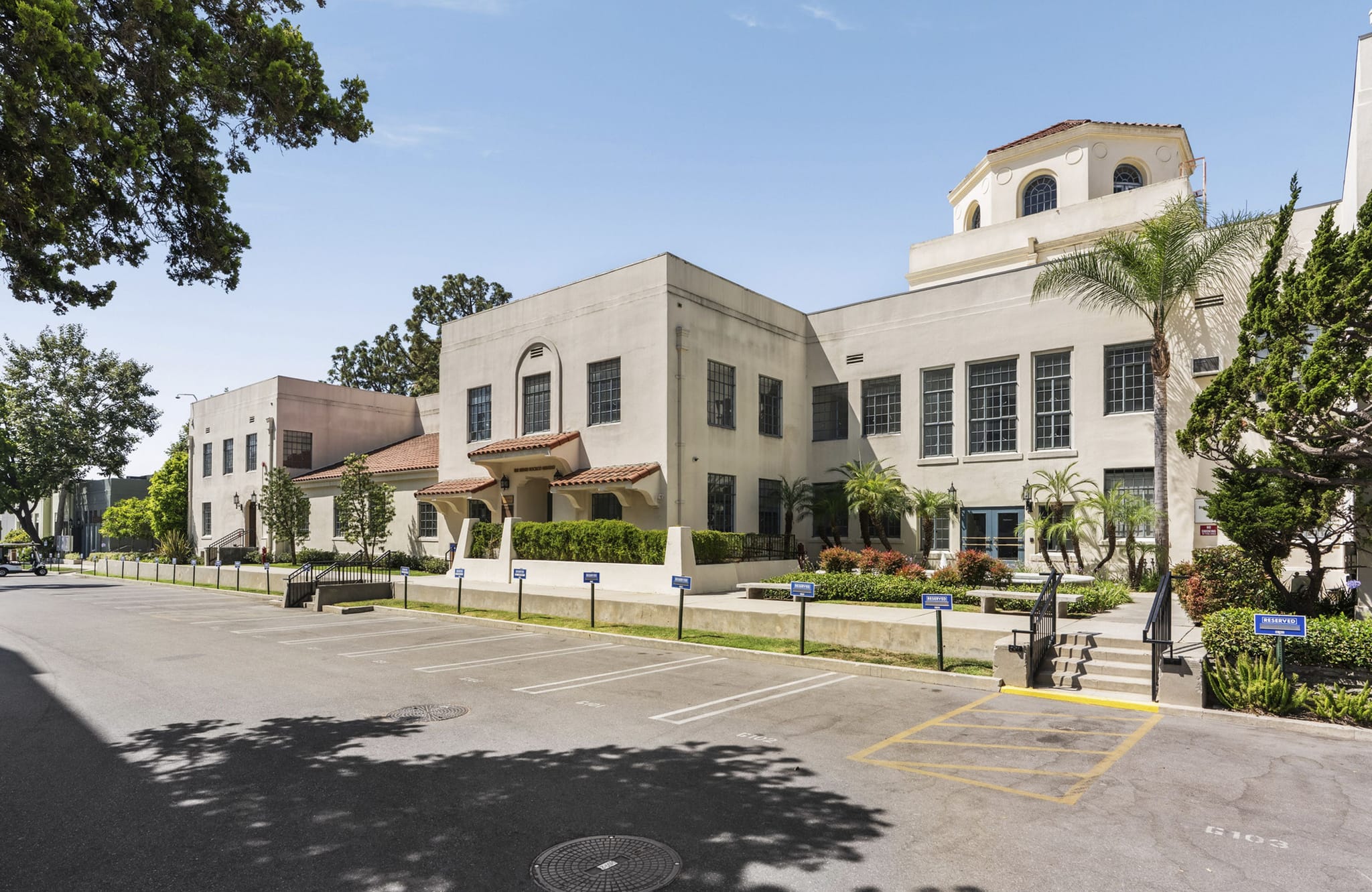 A large, light-colored building in Spanish Colonial Revival style with a domed tower, seen from an empty parking lot on a sunny day.
