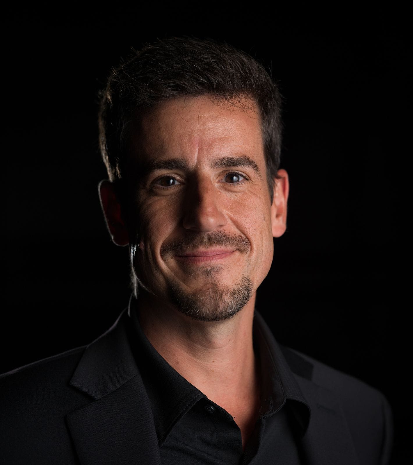 Smiling man with dark hair, mustache, and goatee, wearing a black suit and shirt against a dark background.