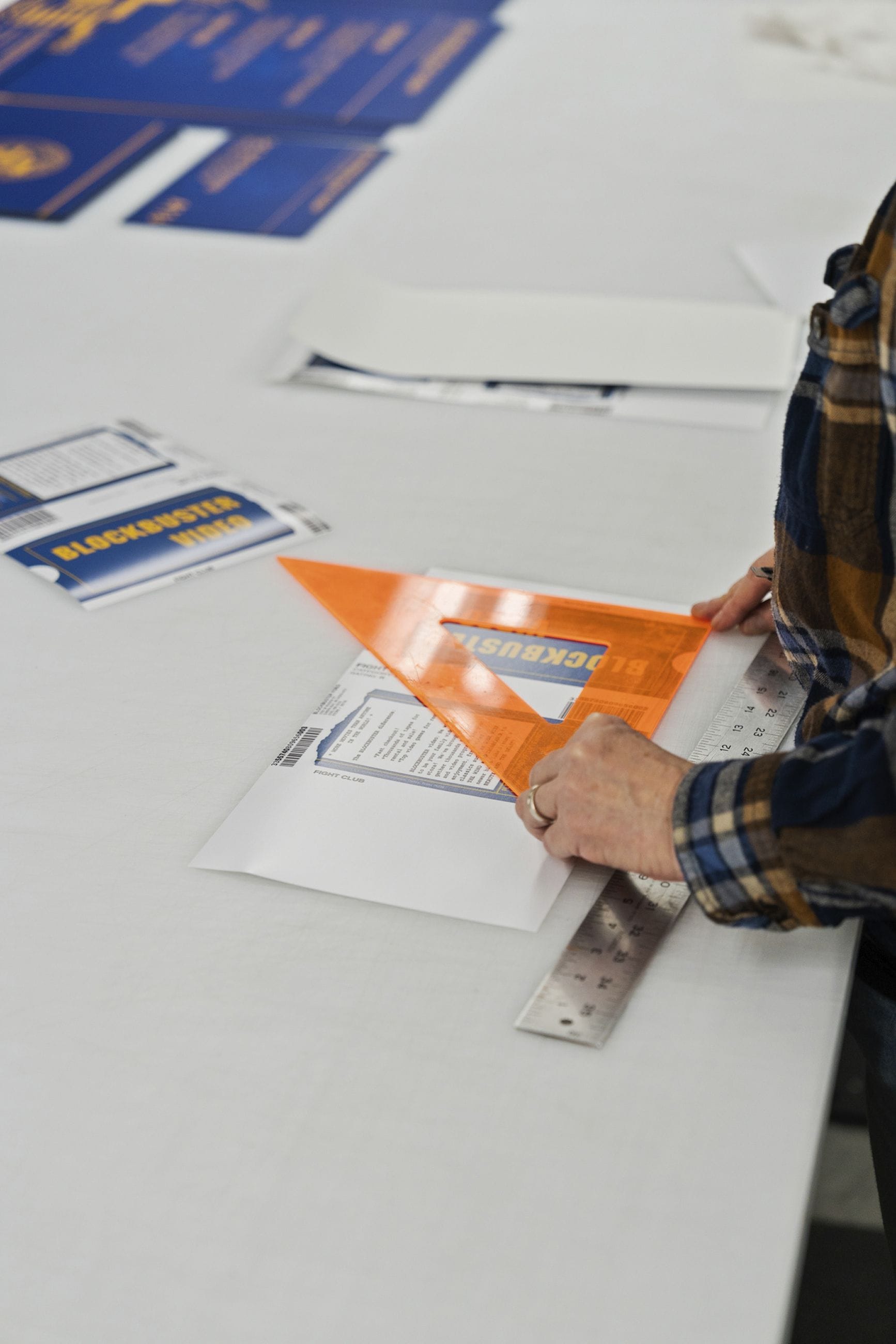 A person wearing a plaid shirt uses an orange triangular ruler and a metal straightedge on Blockbuster Video paperwork.