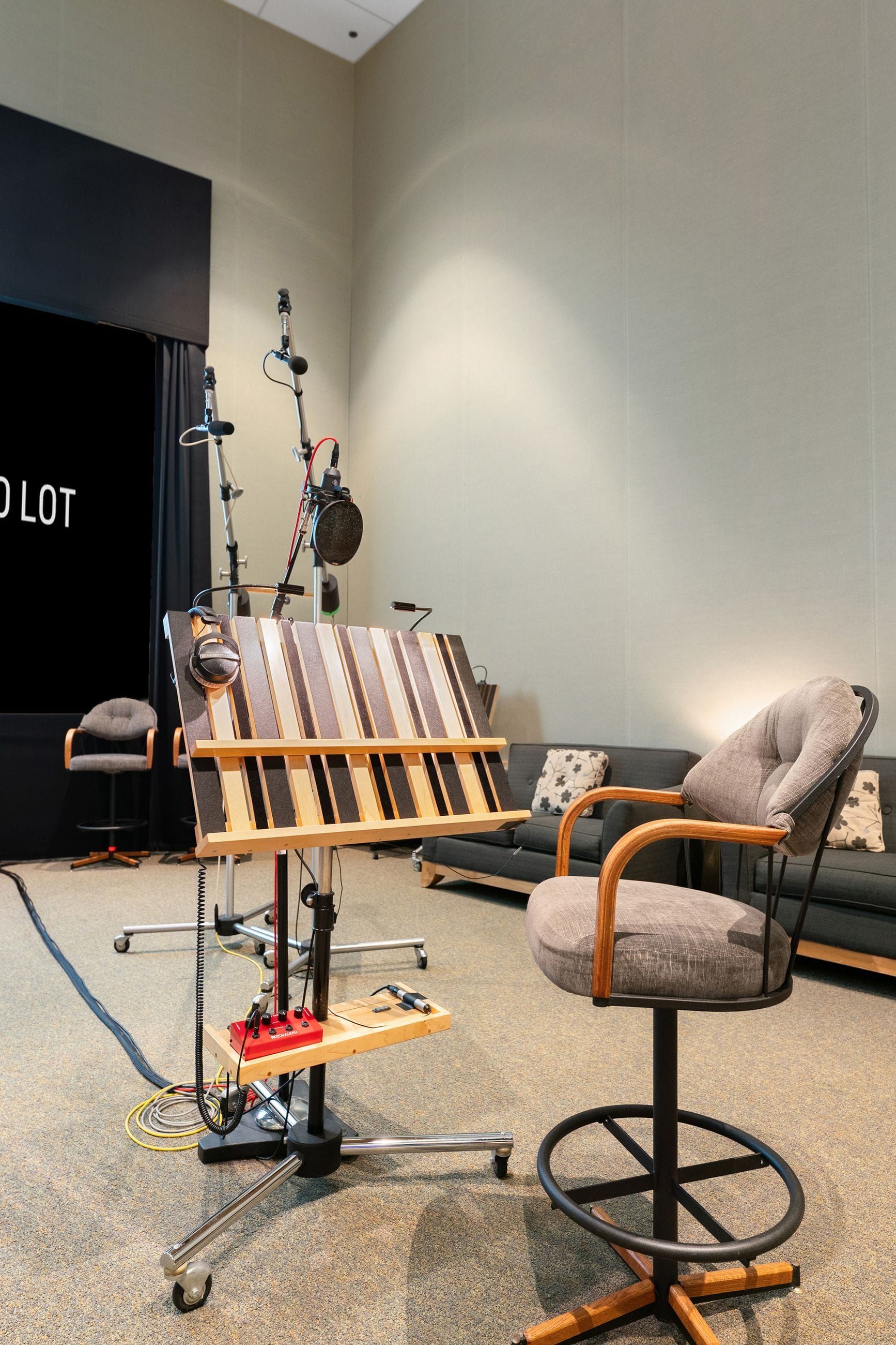 A modern recording studio setup with microphones, a large screen, a distinctive wooden music stand with headphones, and a gray upholstered chair.