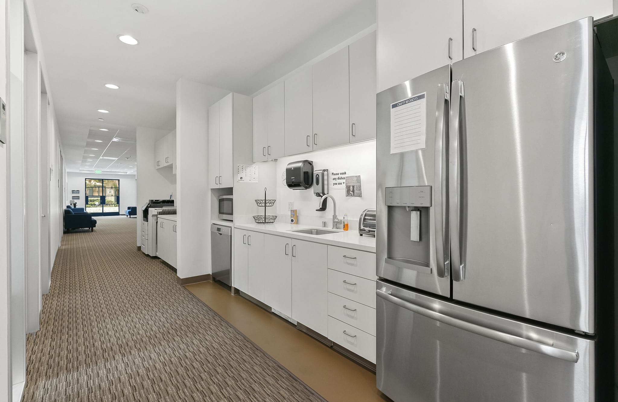 Modern office kitchen with white cabinets, stainless steel appliances, and a carpeted hallway.