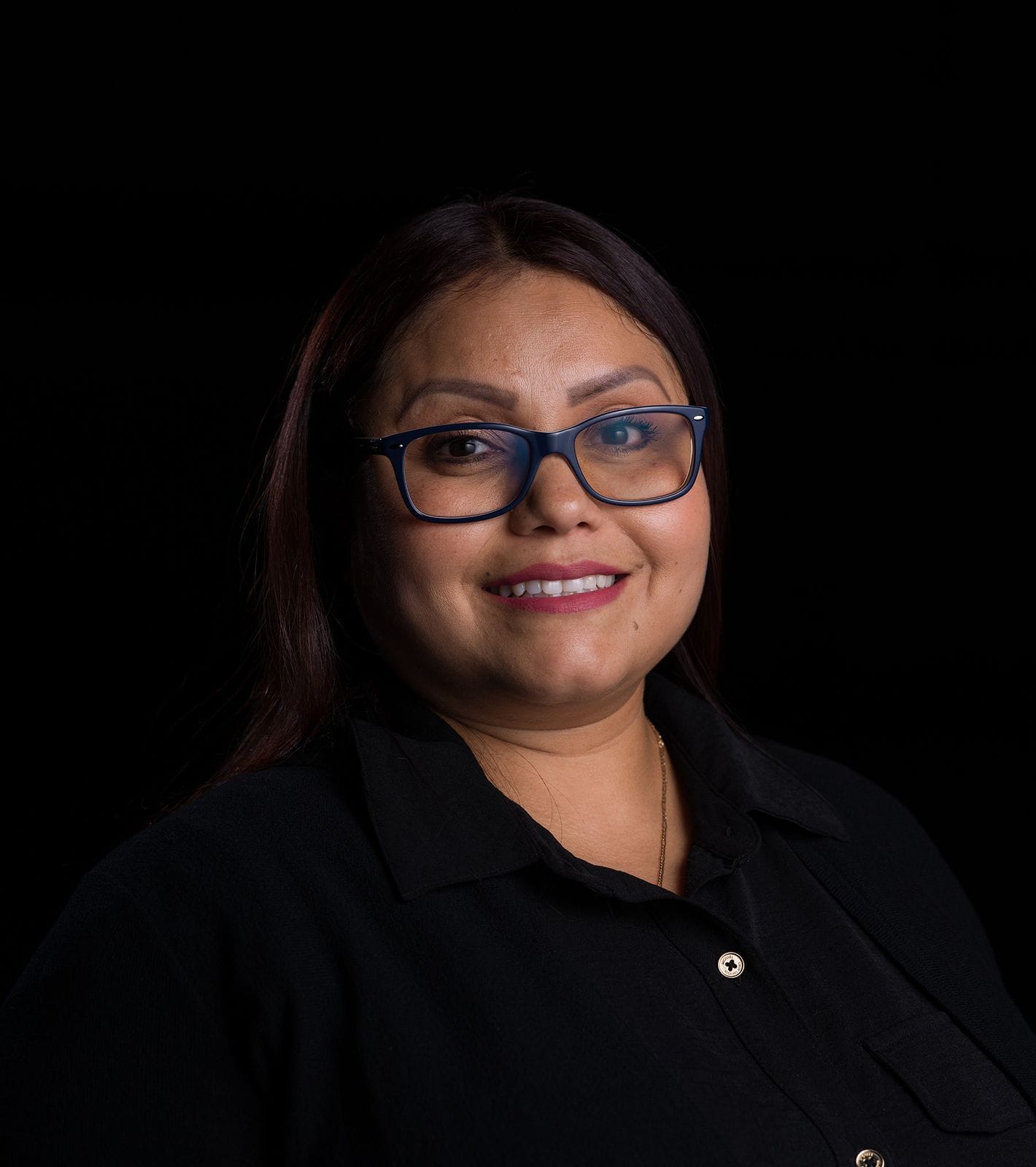 A smiling woman with dark hair and glasses wears a black shirt against a dark background.