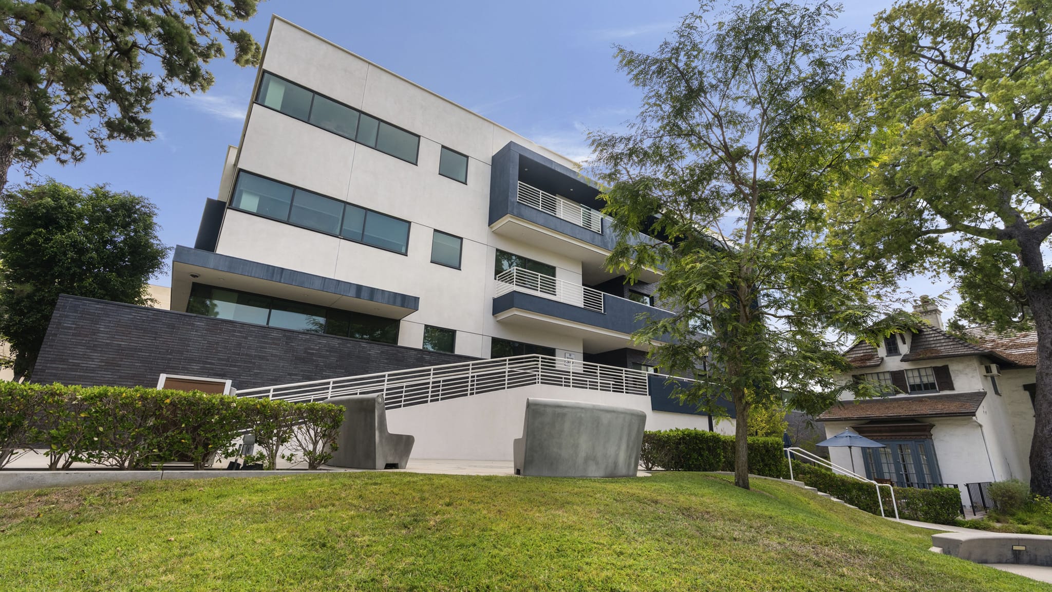 A modern white and blue building on a grassy slope, with an older white house to the right.