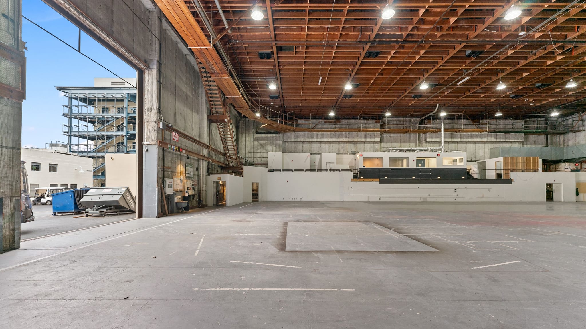 Large empty industrial warehouse with a high wooden ceiling, concrete floor, an open loading bay to an outdoor street, and a raised platform structure inside.