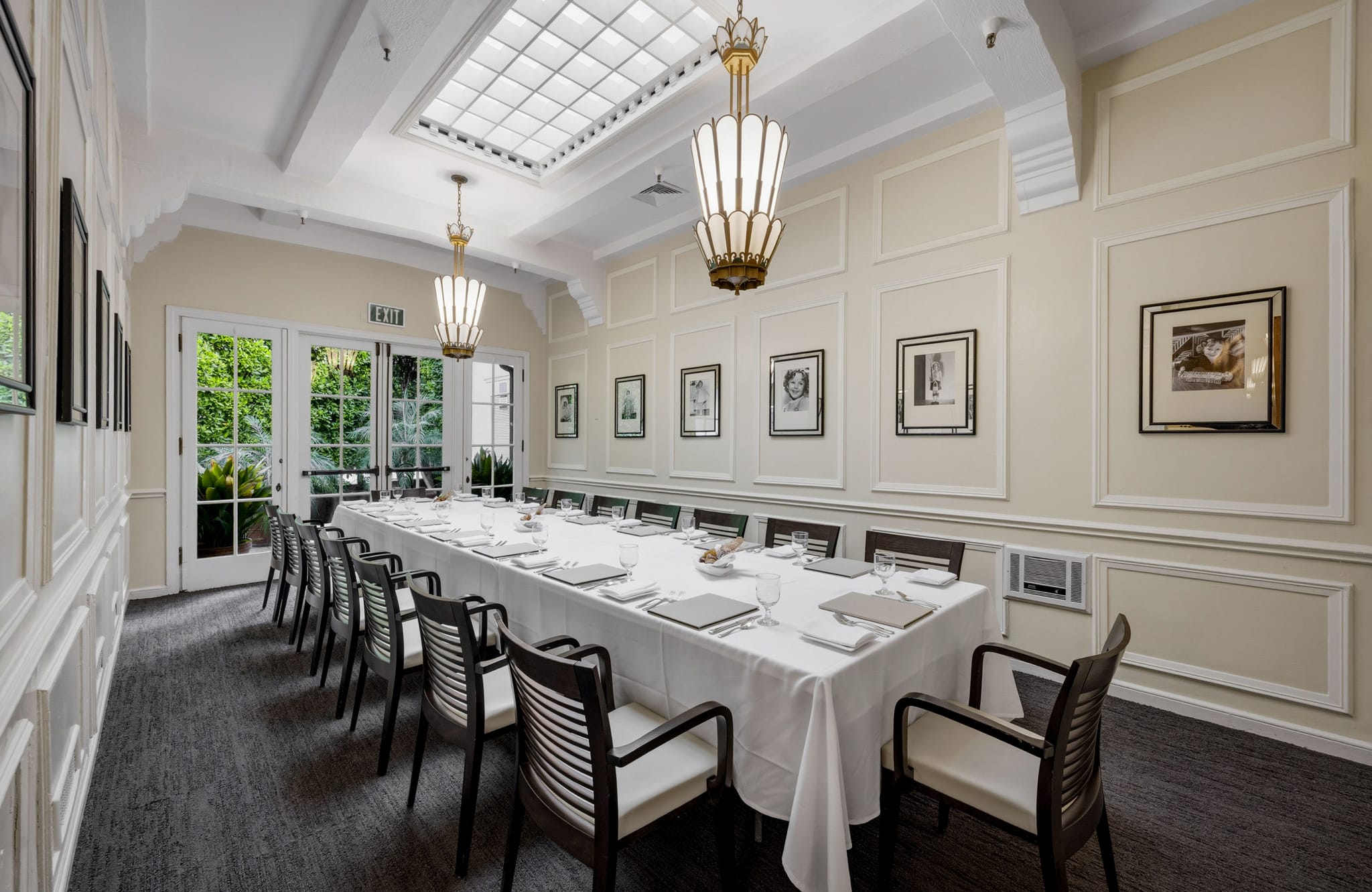 A private dining room with a long white-linened table, chairs, framed art on cream walls, chandeliers, a skylight, and French doors to a garden.