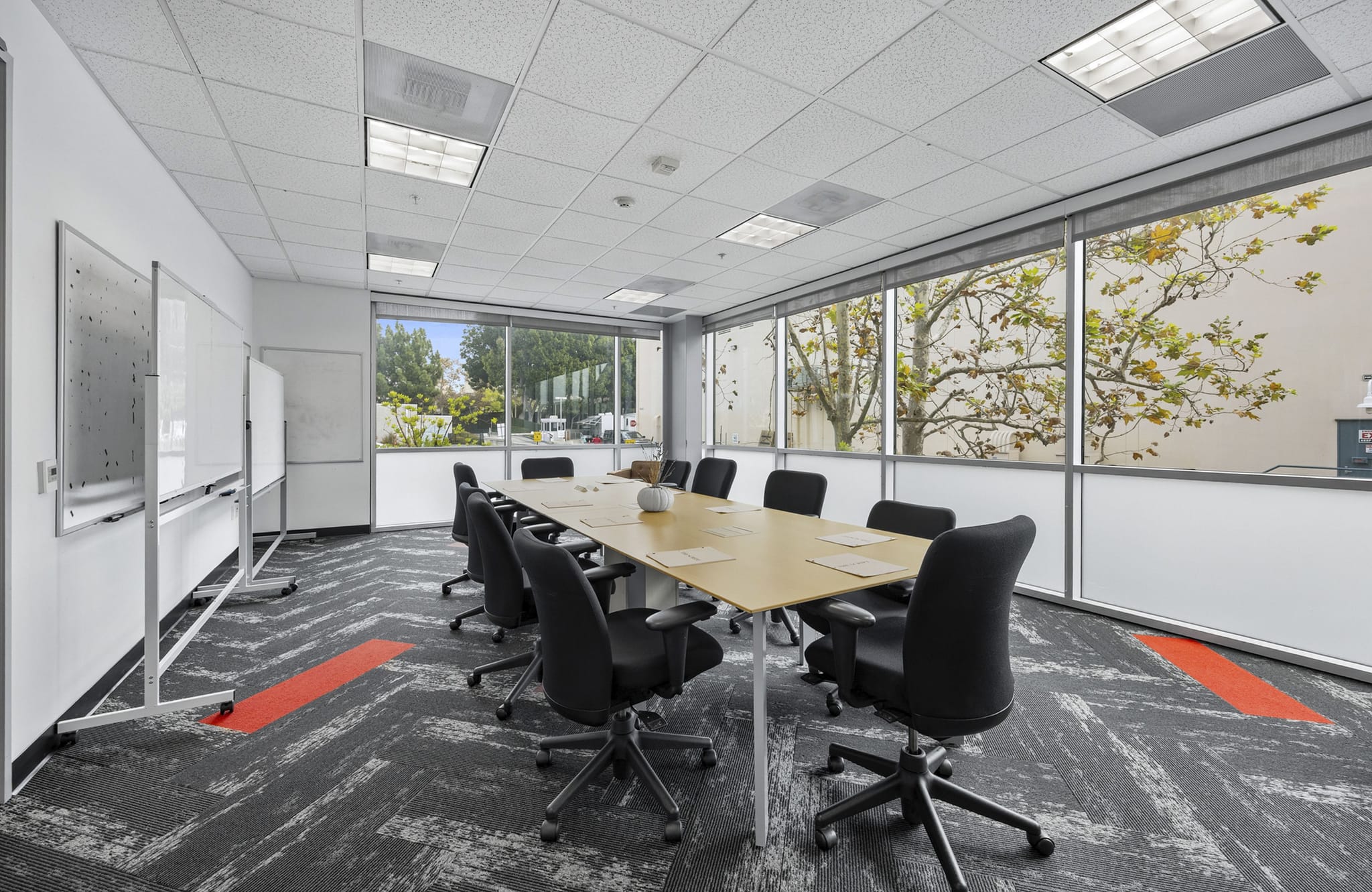 A bright meeting room with a long table, black chairs, whiteboards, and large windows looking out at trees. The floor has a grey patterned carpet with red stripes.