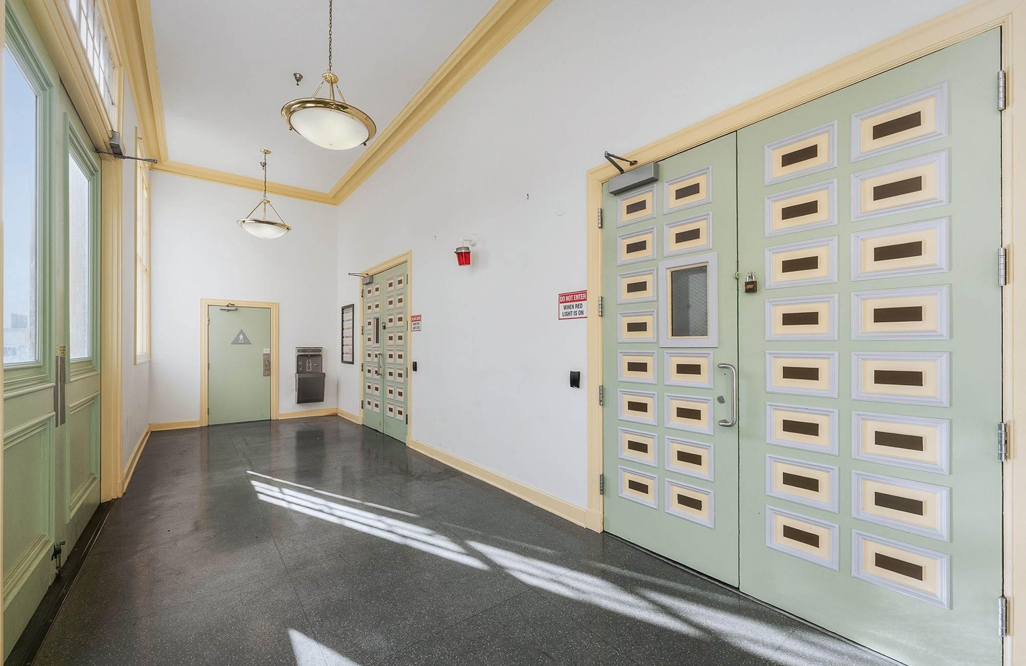 A bright hallway with light green doors featuring decorative rectangular panels, white walls, and a dark floor with sunlight shadows.