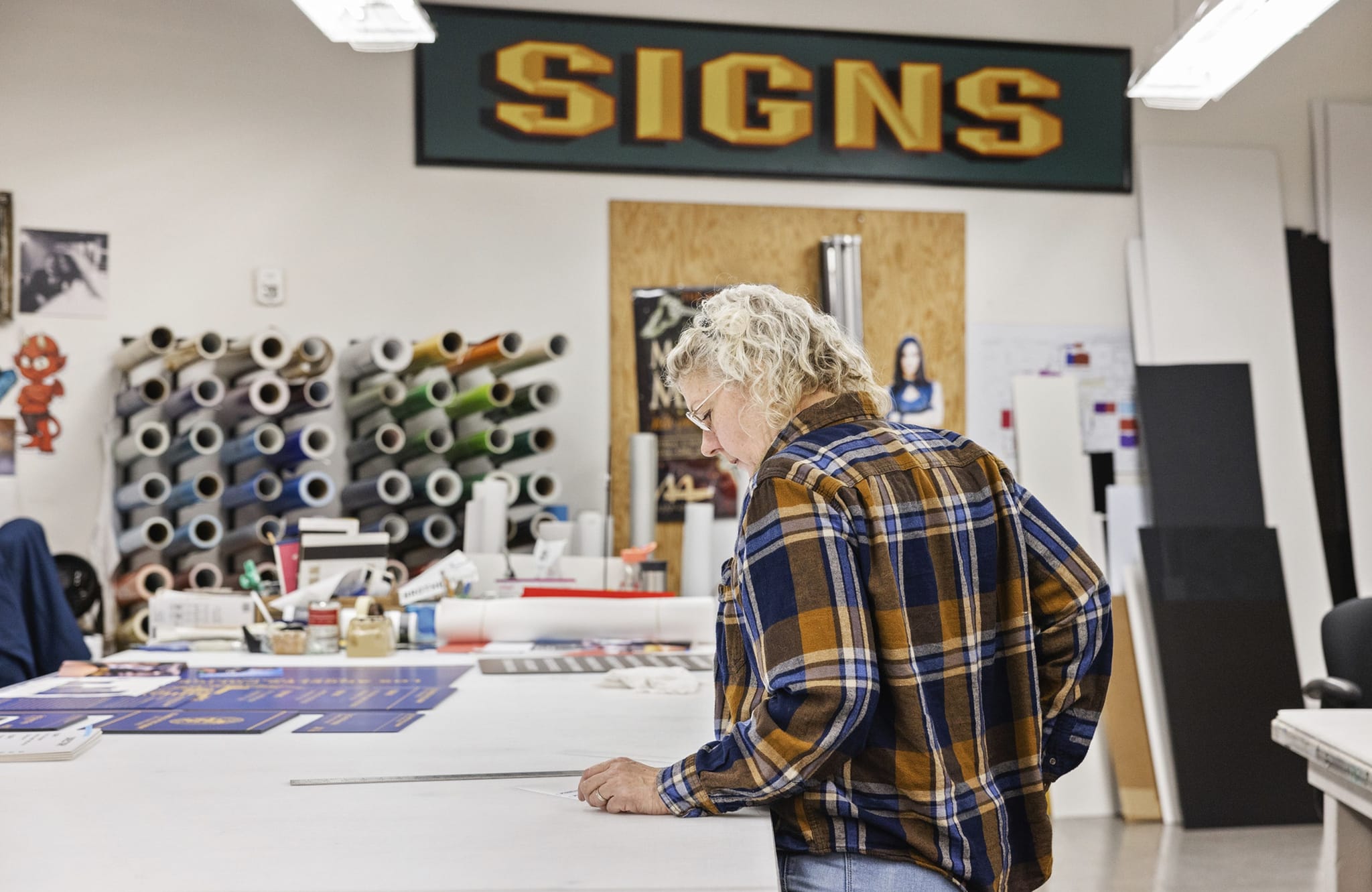 A person in a plaid shirt works at a sign-making table with rolls of material on the wall and a "SIGNS" banner above.