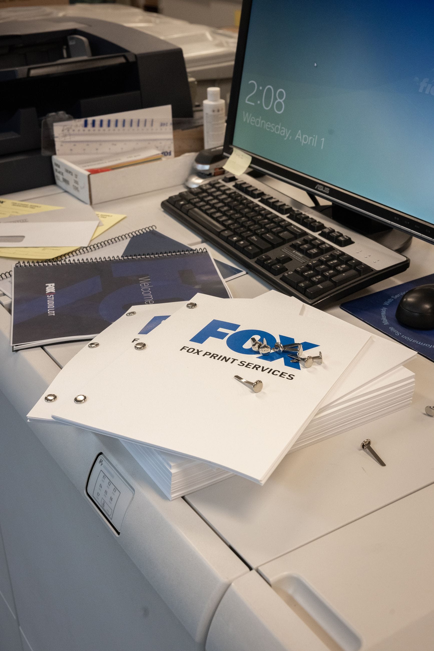 Print shop desk with stacks of "FOX PRINT SERVICES" covers, binding screws, spiral notebooks, a computer displaying "April 1", and a printer.