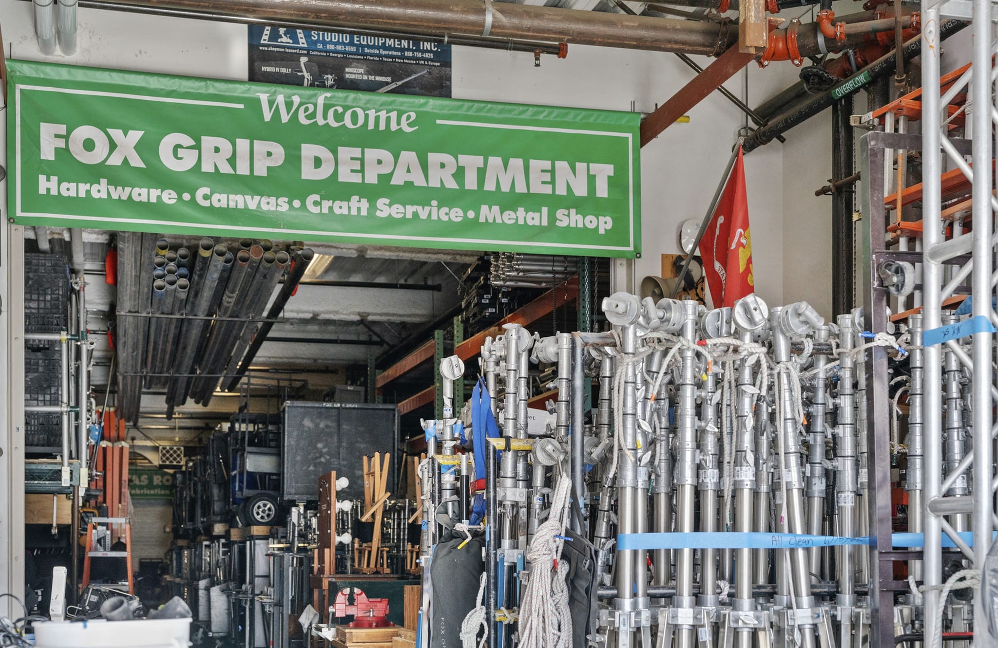 A green "Welcome Fox Grip Department" banner hangs over a warehouse filled with film production grip equipment.