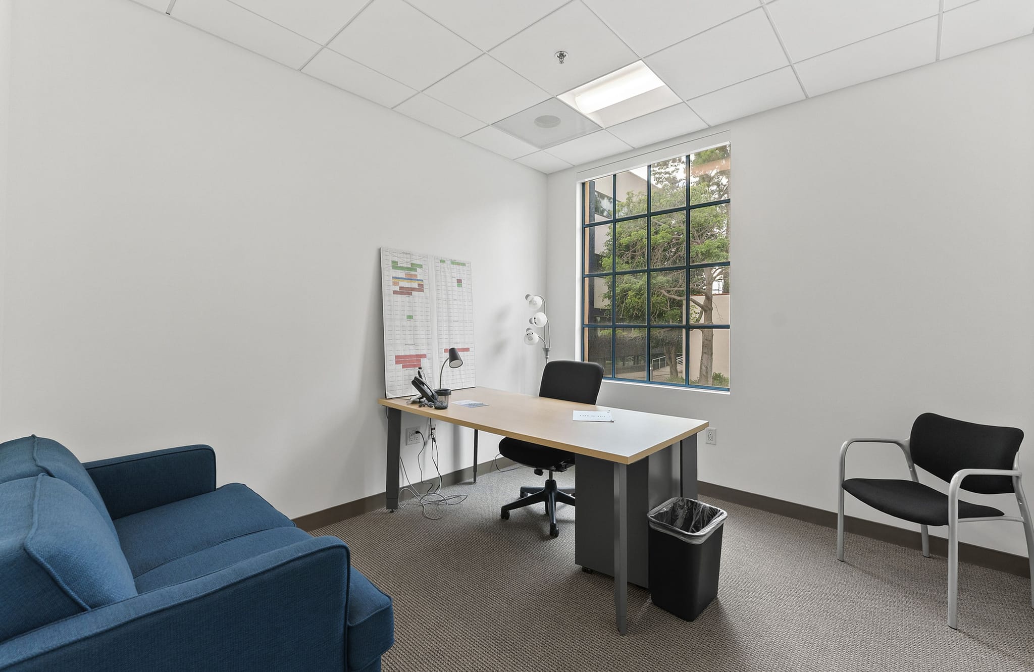 A simple office with a blue sofa, a desk and chair, and a window with a view of trees.