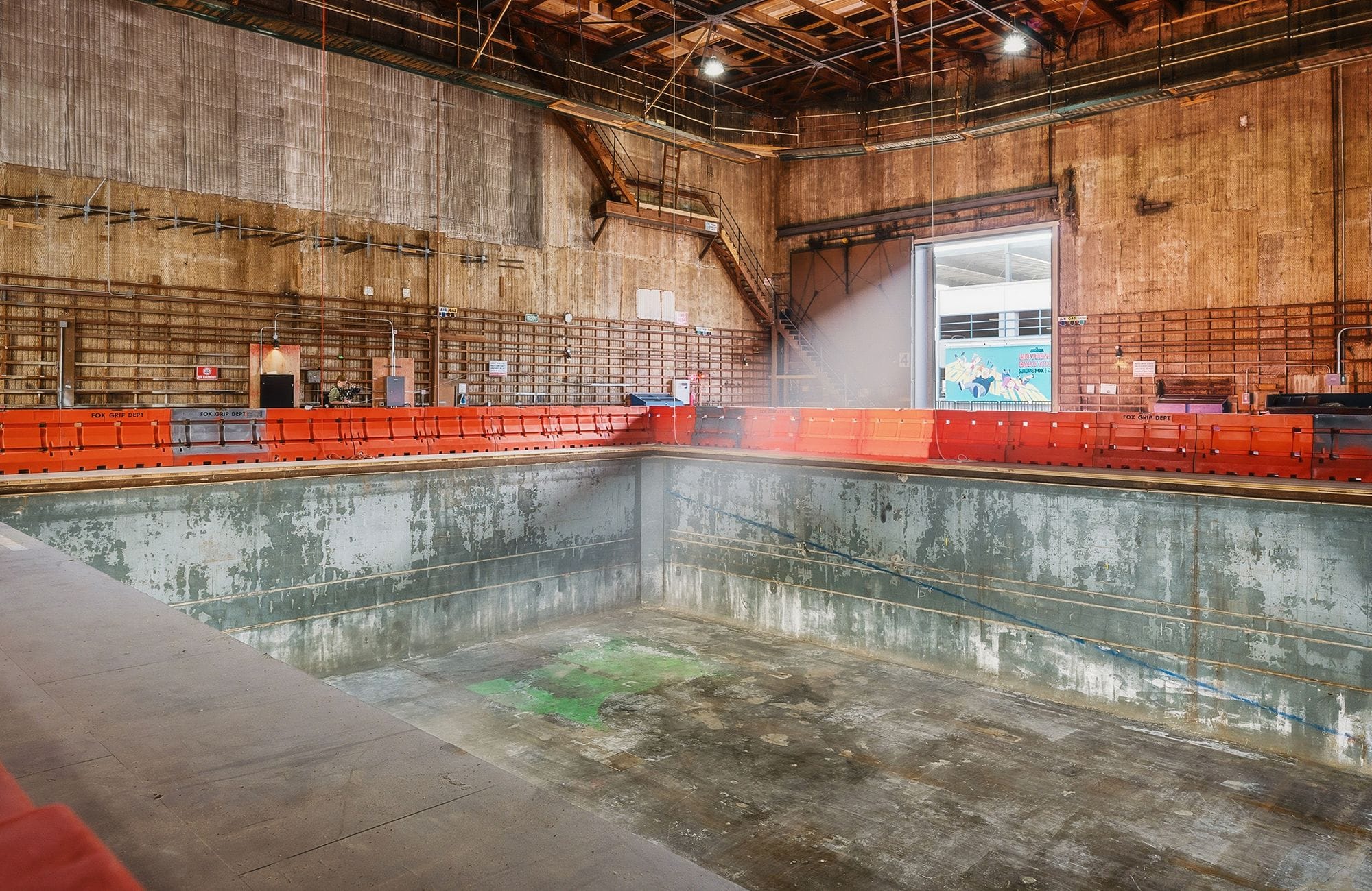 Empty, worn concrete water tank in a large industrial studio with red barriers and light from an open door.